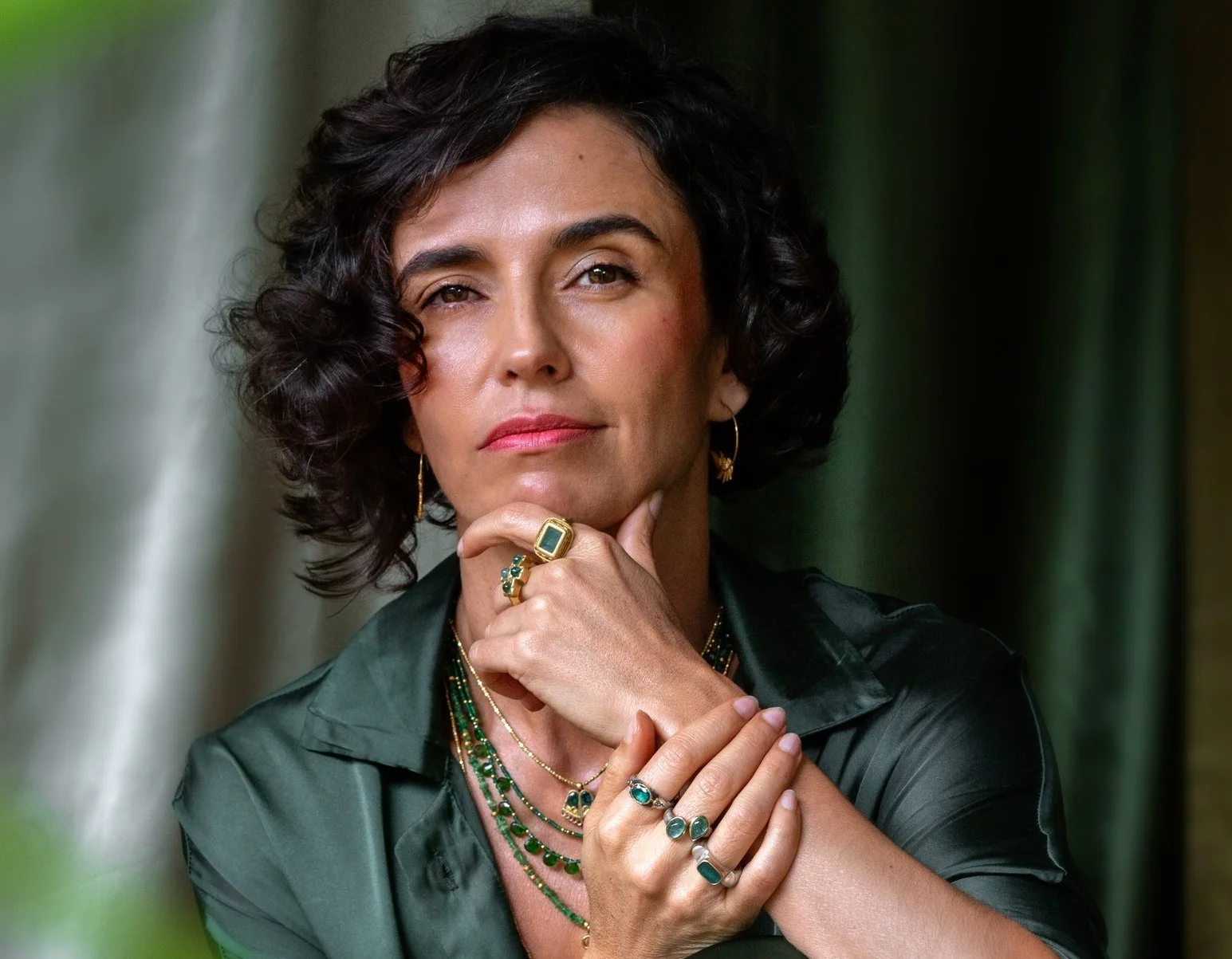 A woman with short, curly dark hair wearing multiple green and gold jewelry, including rings, necklaces, and earrings. She is wearing a dark green satin shirt and posing with her hand on her chin, looking confidently at the camera.
