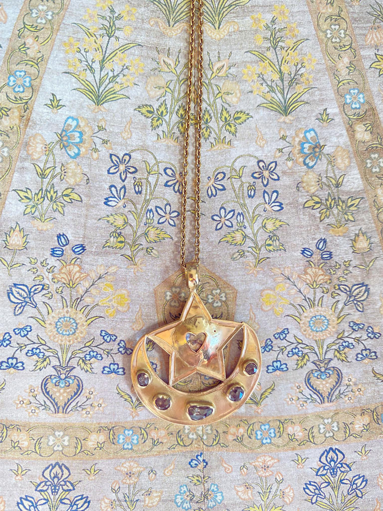 Gold necklace with a moon and star pendant featuring a heart cutout in the star and several small gemstones, hanging over a floral fabric background.