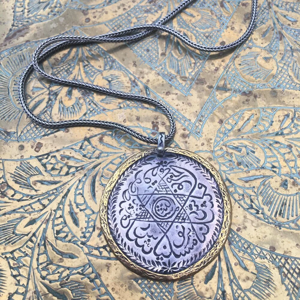 A silver and gold pendant necklace with intricate script and symbols, lying on decorative patterned fabric.