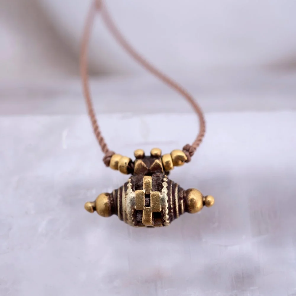 A necklace with a large central pendant and small beads, featuring metallic and dark-colored elements, hanging from a brown string.