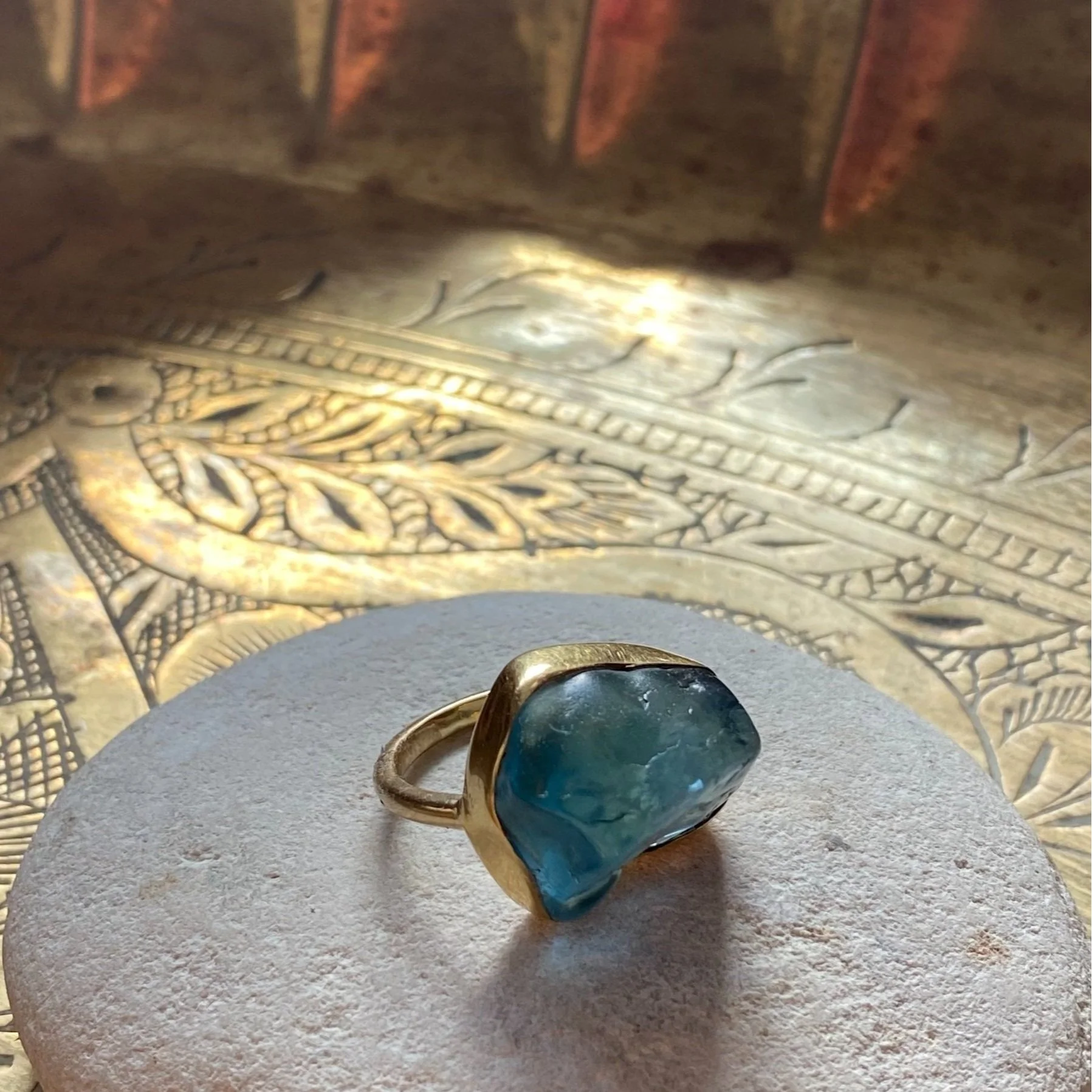 A gold ring with a large, irregularly shaped blue-green gemstone, placed on a round, textured, light-colored surface. In the background, there is intricate, gold-toned decorative metalwork.