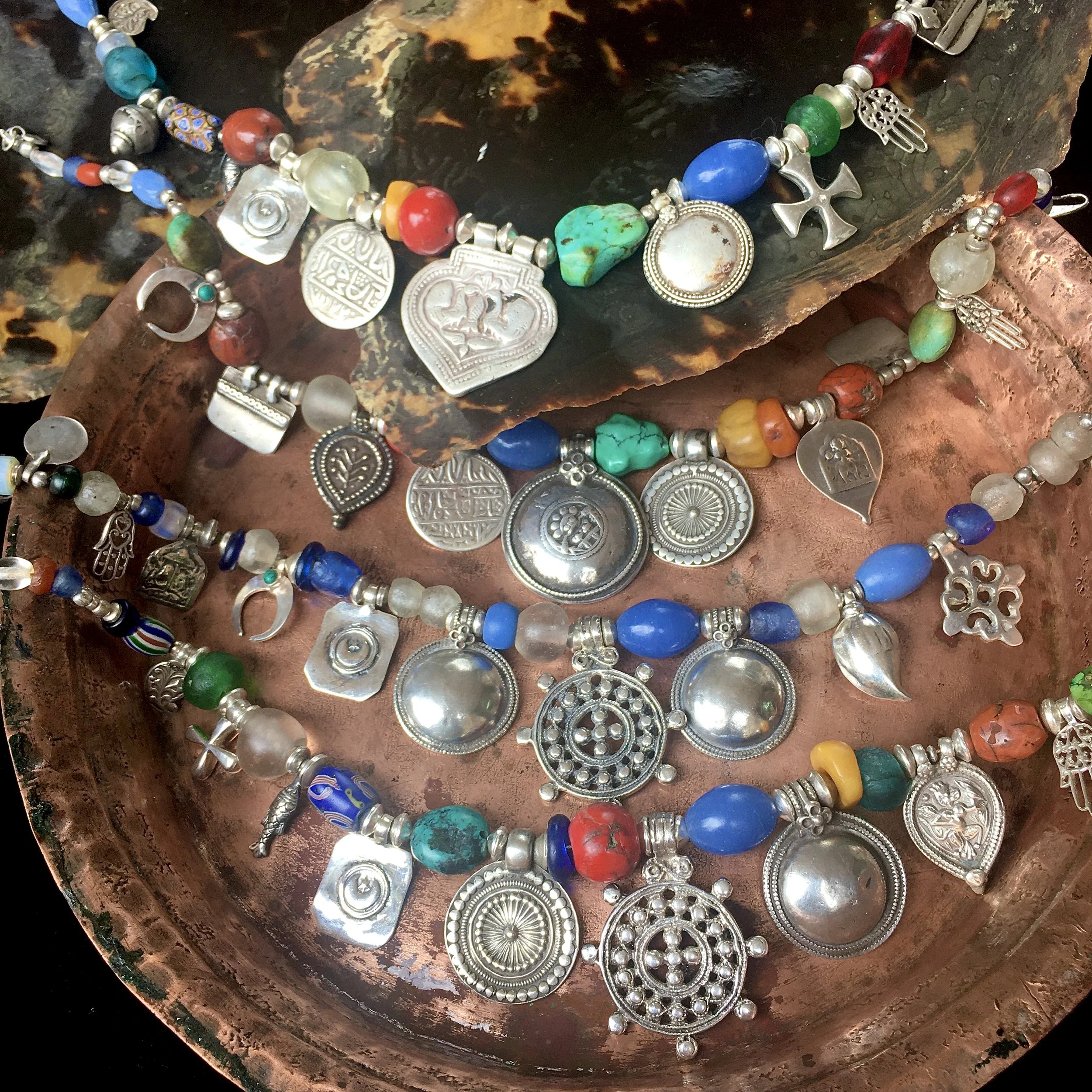 Colorful beaded necklaces with silver charms and pendants, displayed on copper and stone trays.