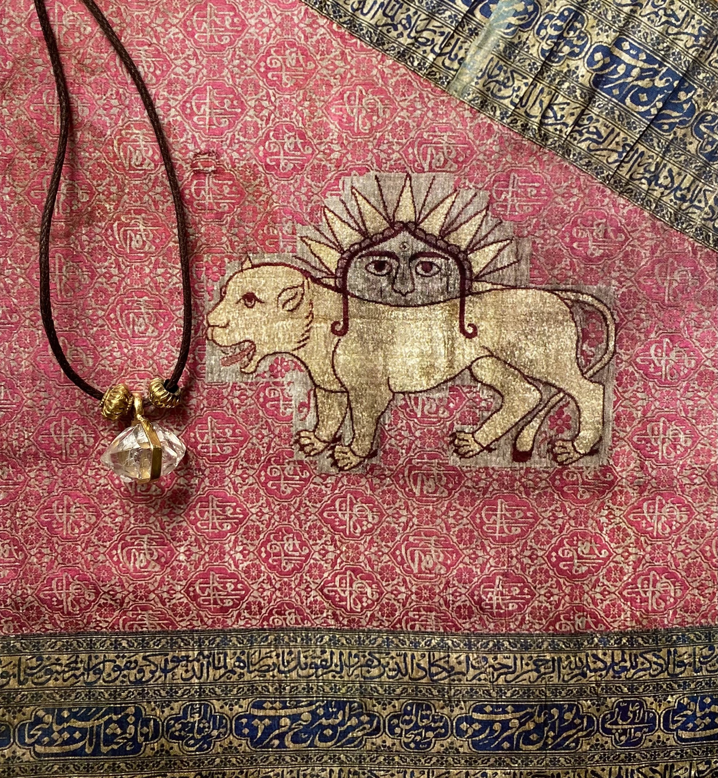 A decorative textile featuring a lion with a human face, six legs, and a sun-like halo. A necklace with a large pendant is placed on the textile.