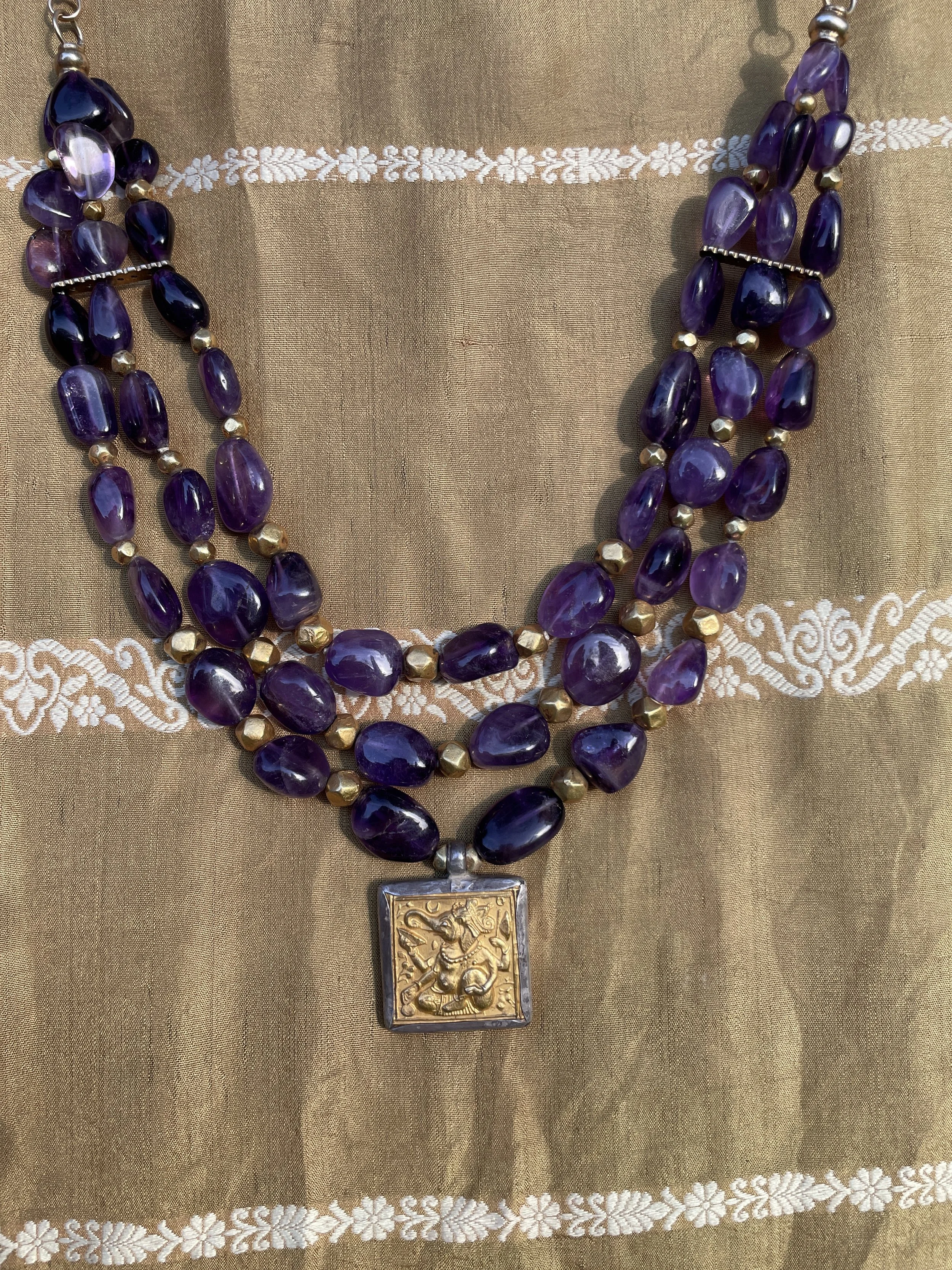 A layered necklace featuring purple oval beads, small gold beads, and a square pendant with a gold embossed image of a figure holding a spear, on a gold fabric background with white embroidered floral patterns.