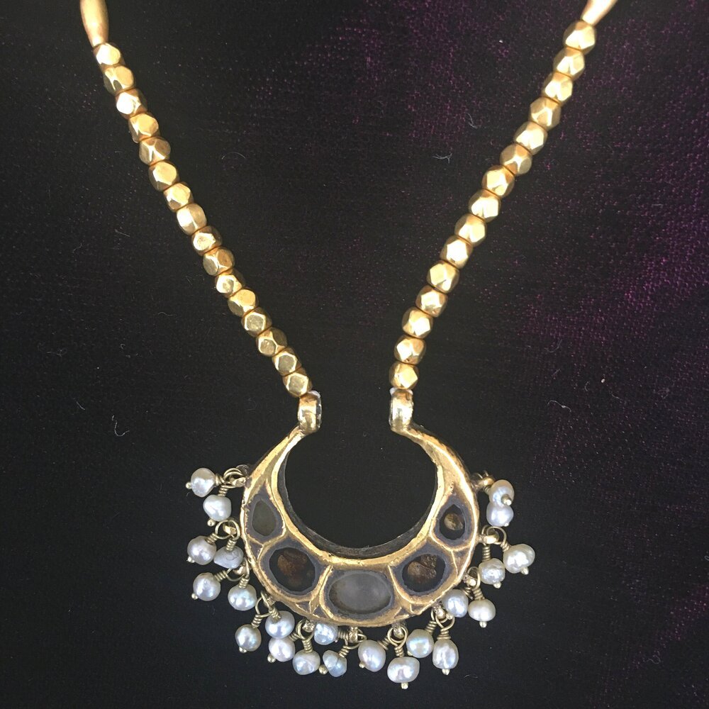 A gold necklace with white beads and a large ornate pendant with pearl accents on a dark fabric background.