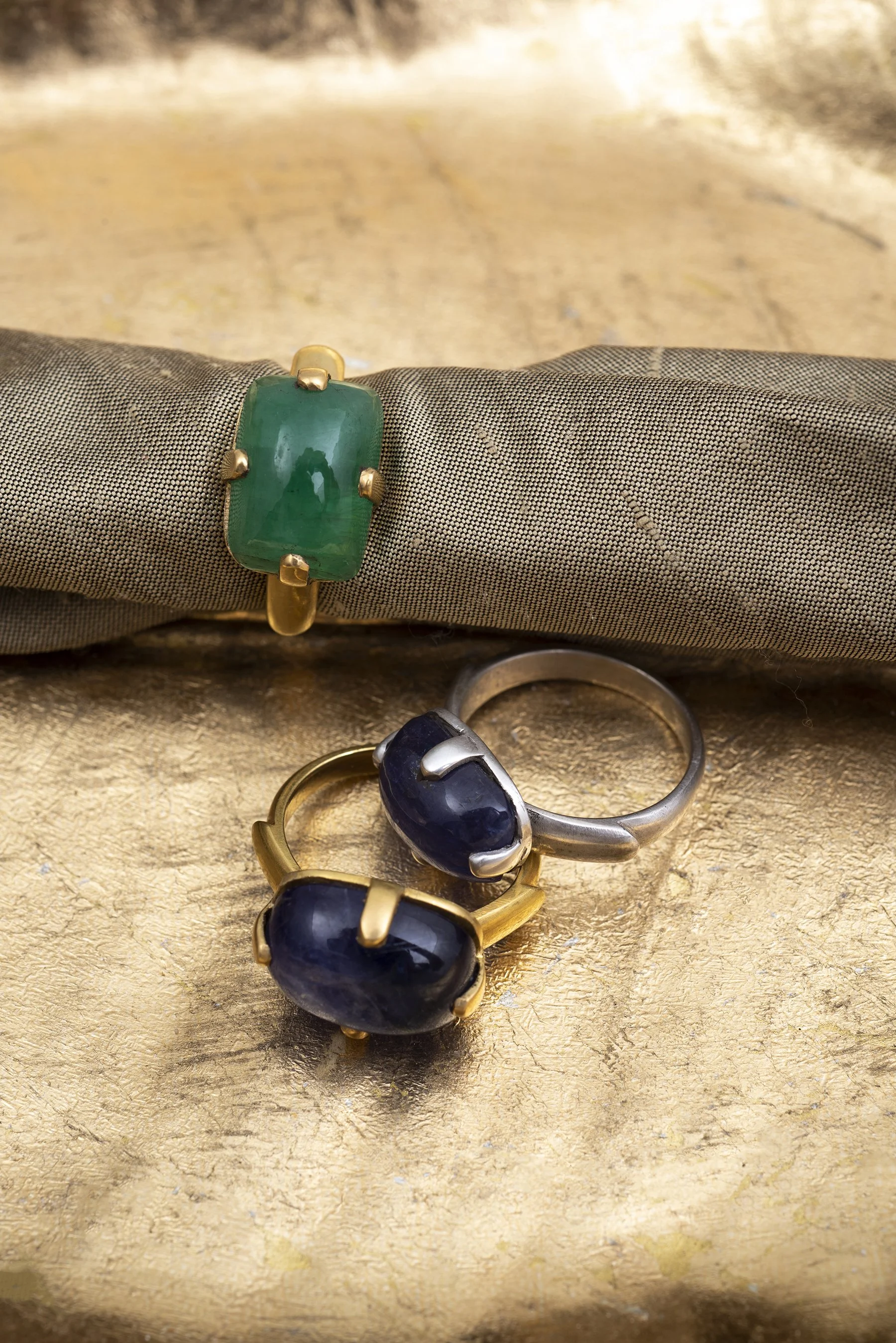 Three rings with large polished stones, a green one with a gold band on a fabric surface, and two with dark blue stones, one with a silver band and one with a gold band, on a textured gold surface.