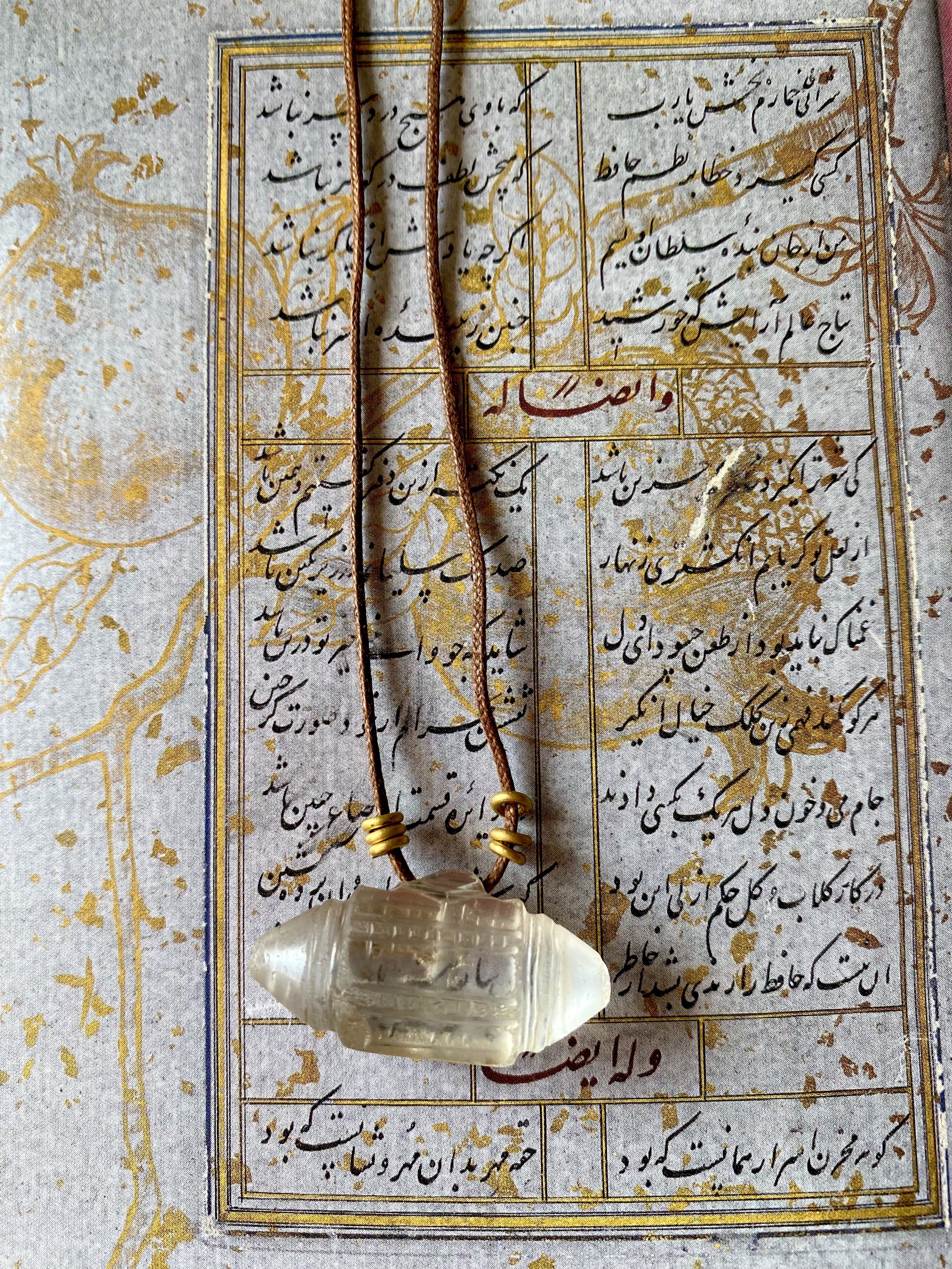 A necklace with a carved white pendant rests on a piece of paper with Persian calligraphy in black ink and gold accents.