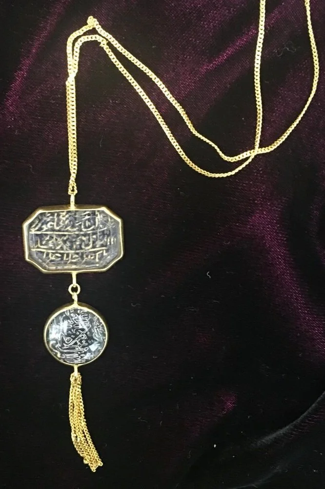 Gold necklace with two pendants containing Arabic script, one rectangular and one circular, with gold tassels at the bottom, lying on a dark fabric background.