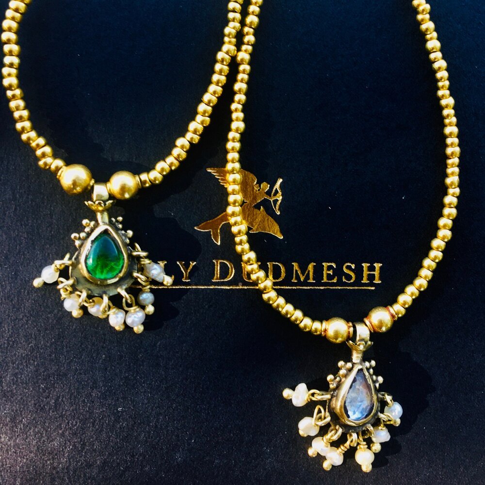 Two gold necklaces with large teardrop-shaped pendants, one with green gemstone and the other with clear gemstone, surrounded by small pearls, placed on a black surface with the brand name "LILY DEDMESH" and a bird logo.