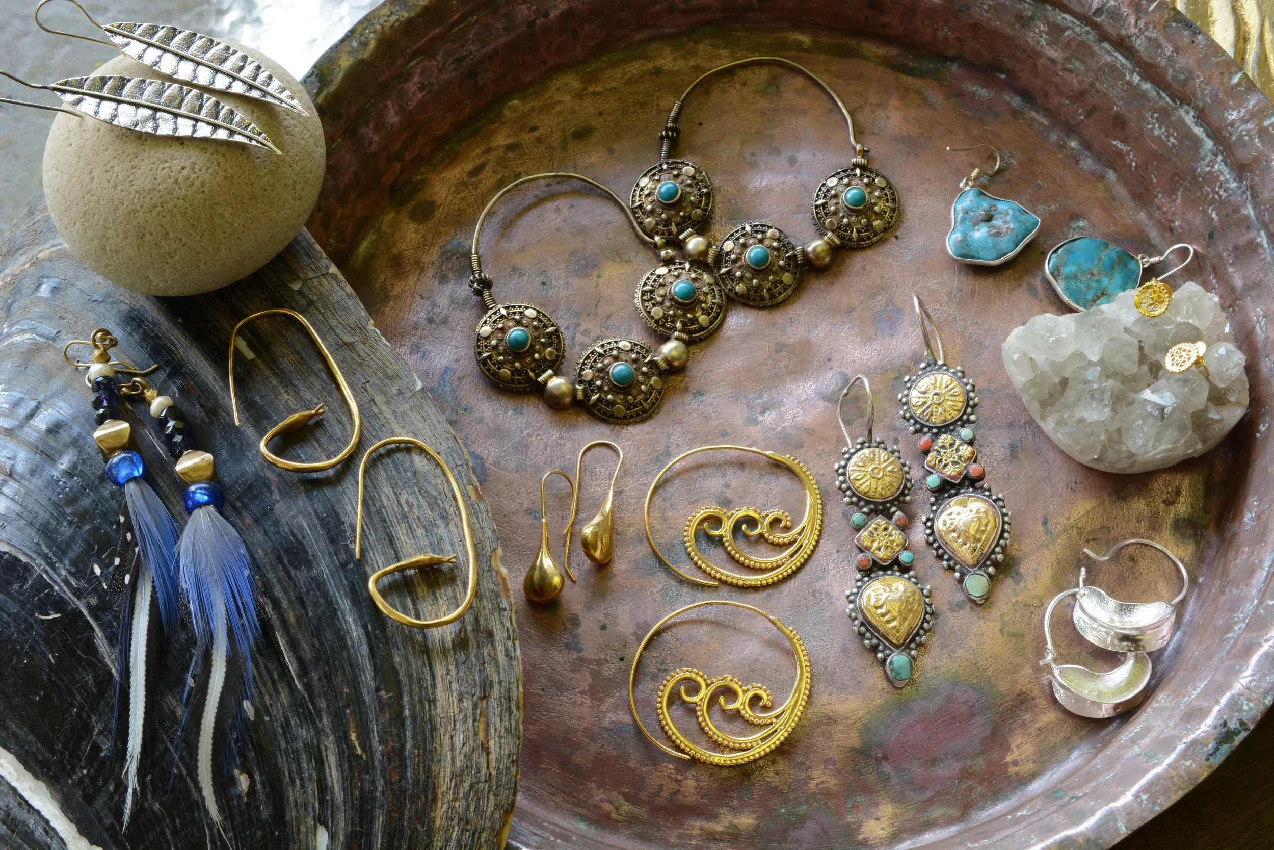 Collection of assorted jewelry including earrings, necklaces, and rings on a rustic metal tray and a wooden surface, featuring turquoise, gold, silver, and bead designs.