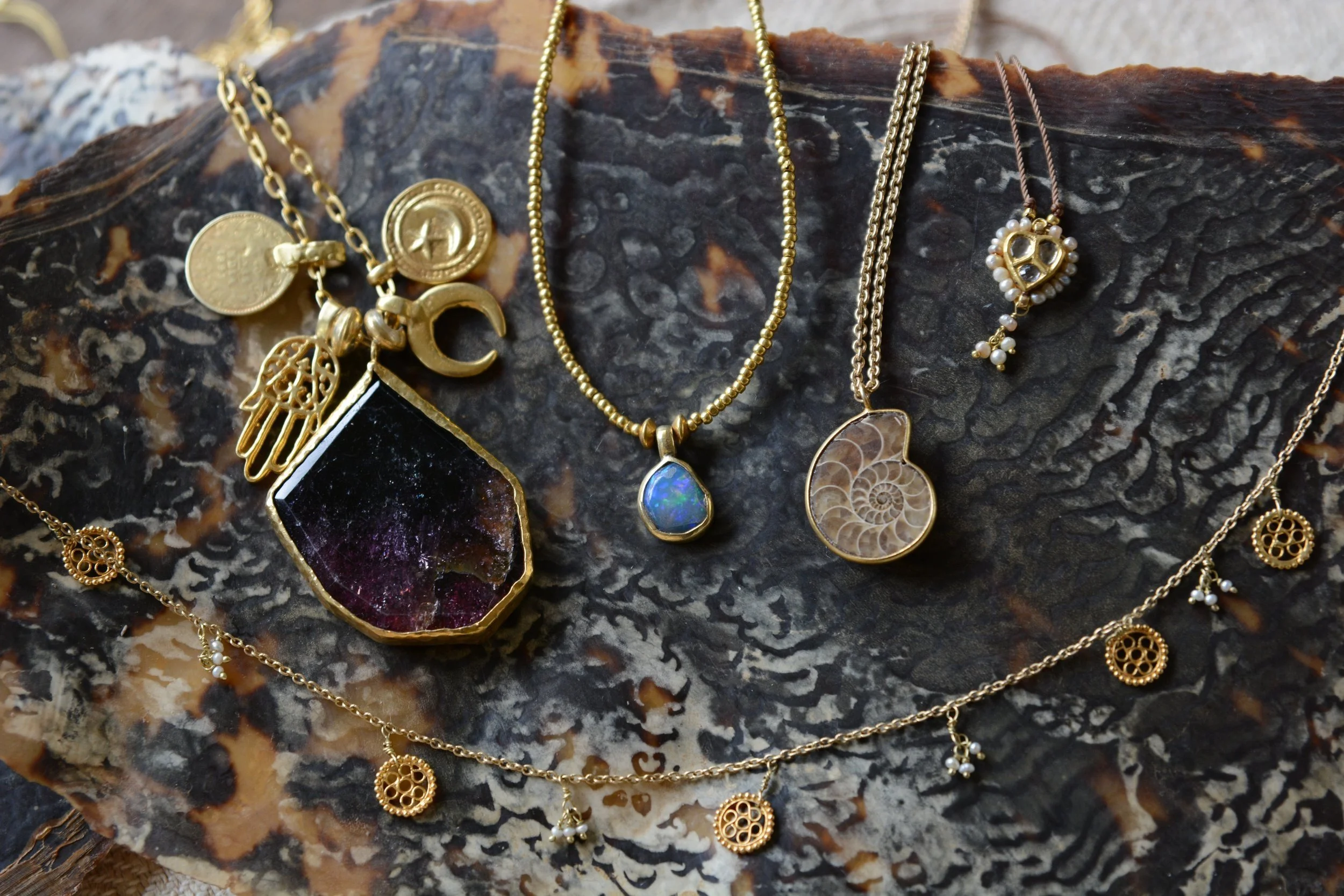 Several gold necklaces with various charms and pendants, placed on a dark, textured rock surface. The jewelry includes a pendant with a dark purple and black stone, a turquoise stone pendant, a shell fossil pendant, and decorative charms like a Hamsa hand, a peace sign, a heart outlined with small pearls, and small round charms with intricate designs and tiny pearl accents.