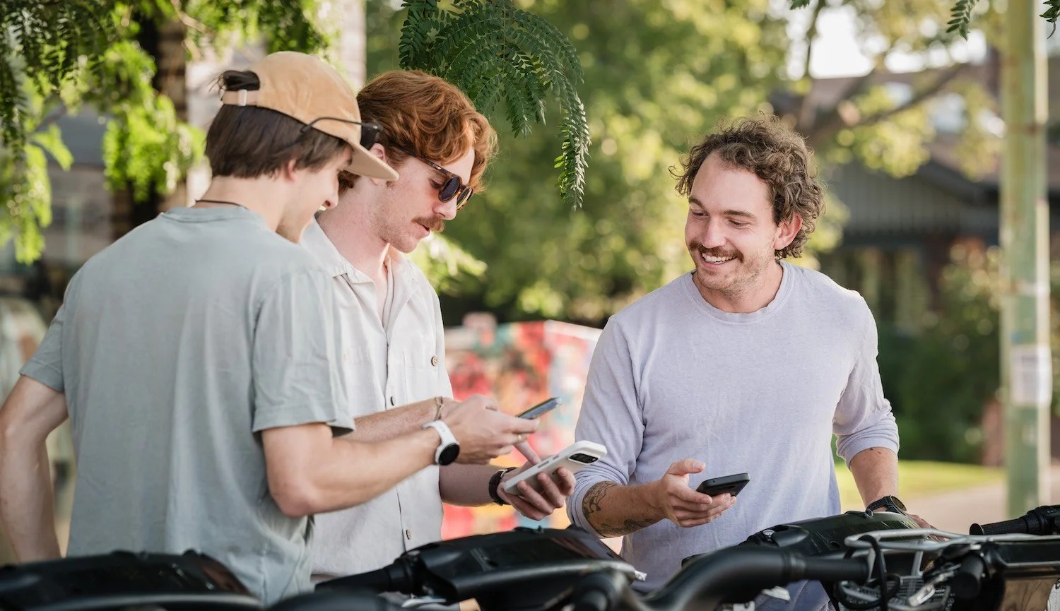Four men standing outdoors under trees, smiling and looking at their smartphones, with bicycles in the foreground.