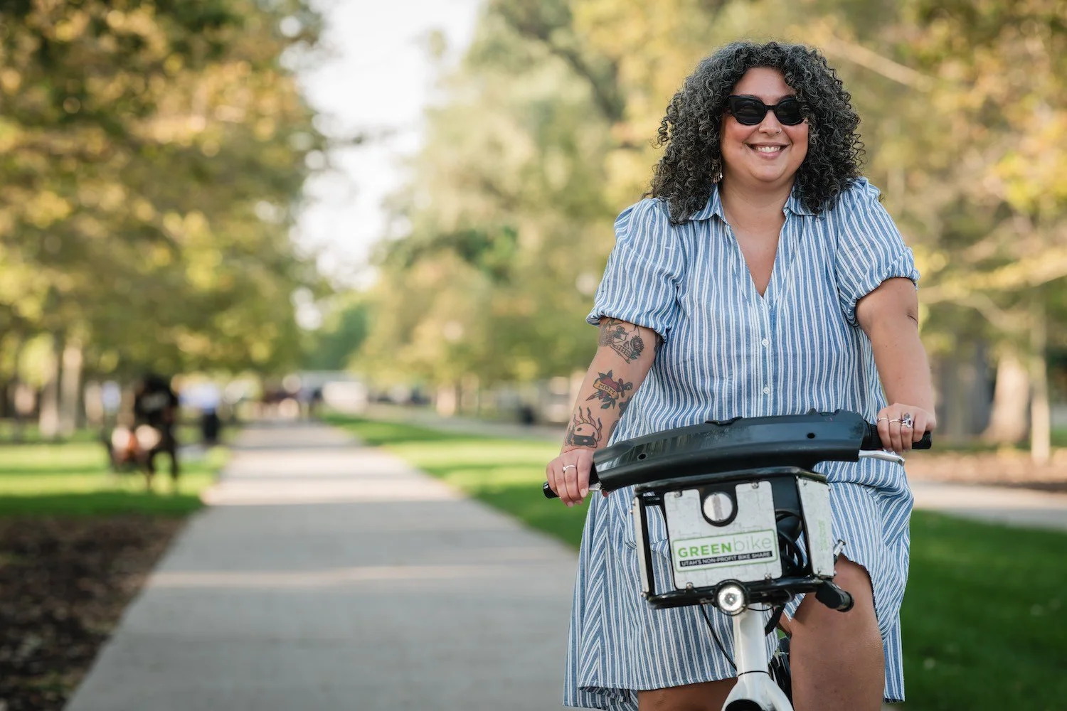 A woman with curly hair, sunglasses, and a striped dress riding a bike along a tree-lined path in a park.