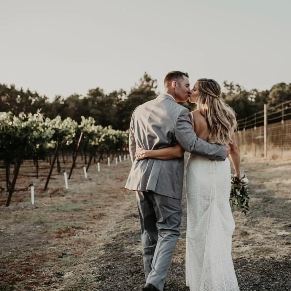 A couple in wedding attire sharing a kiss in a vineyard during sunset.