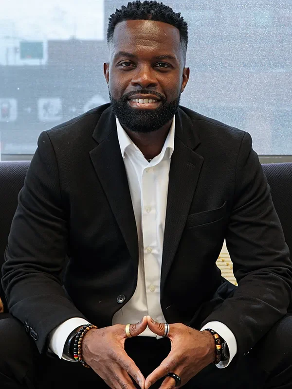 Homme d'affaires afro-américain souriant, en costume noir et chemise blanche, assis à un bureau avec un fond de fenêtre.