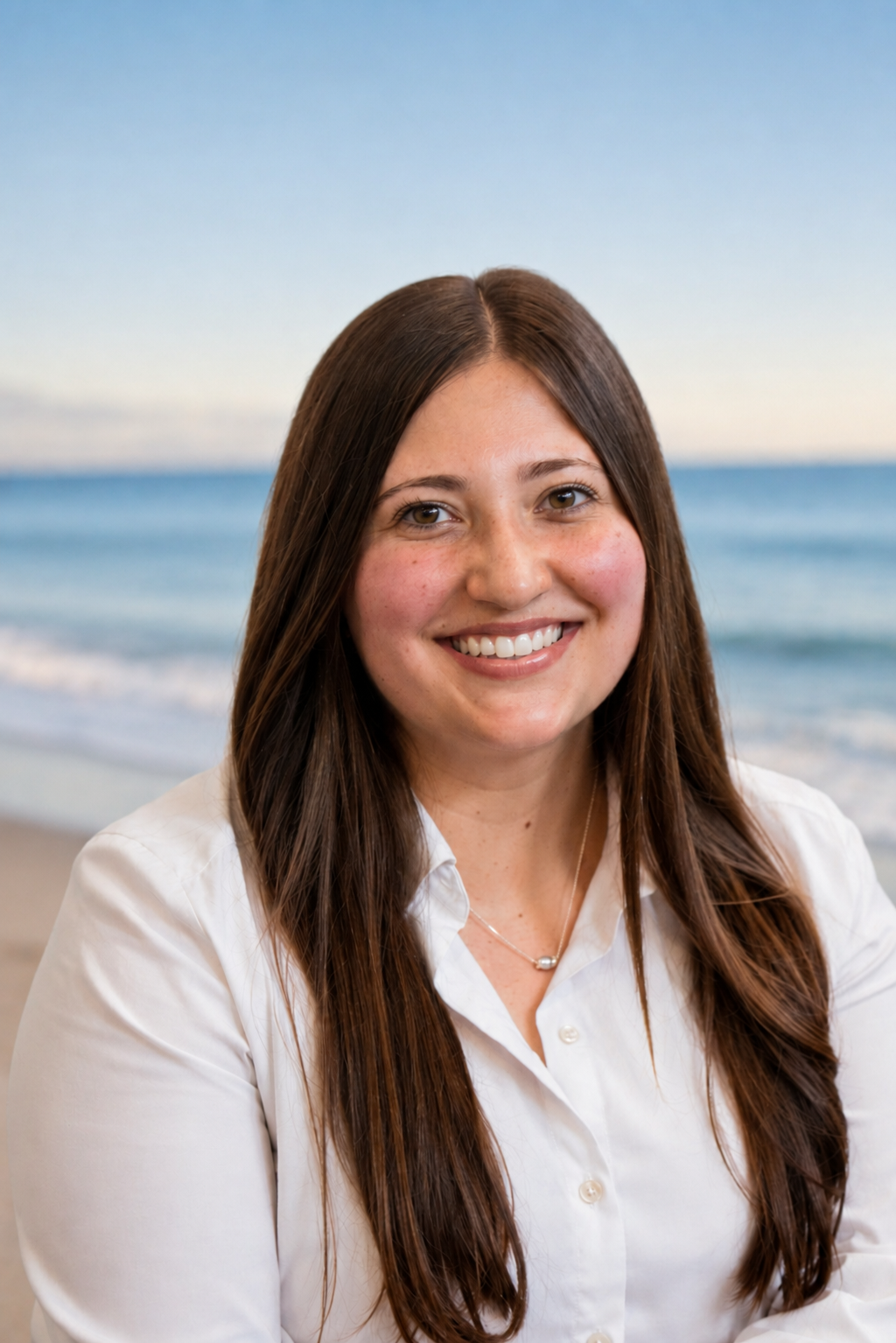 A young woman with long brown hair smiling at the camera on a beach with the ocean and sky in the background.