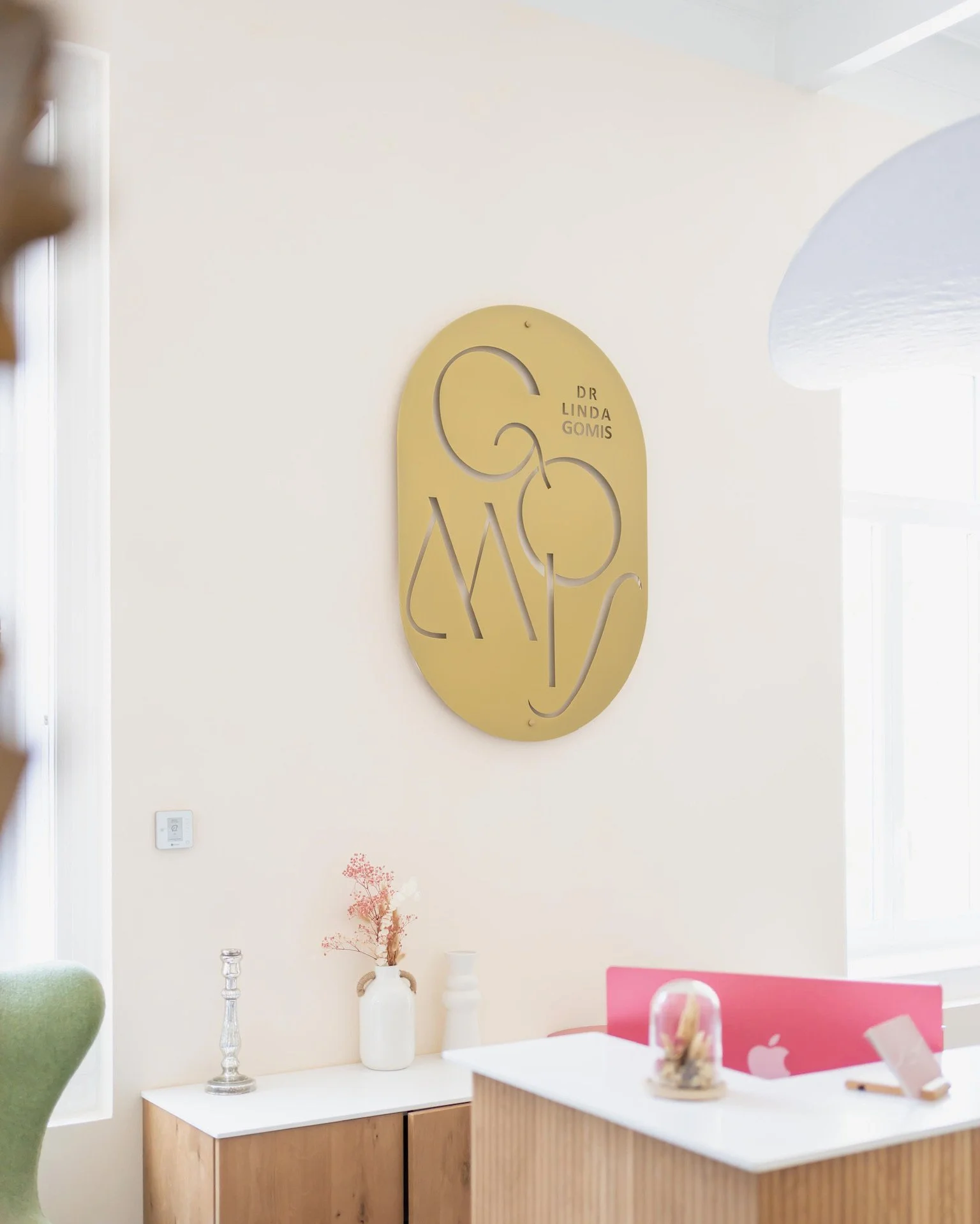 Une décoration murale en métal doré avec le texte 'GMO' et 'Dr Linda Gomis' sur un mur blanc. Au premier plan, un bureau blanc avec un visage en verre avec de petites fleurs à l'intérieur, une tablette rose avec un logo Apple, et des objets décoratifs blancs. À gauche, un meuble en bois avec deux vases, dont un avec des branches de fleurs séchées.