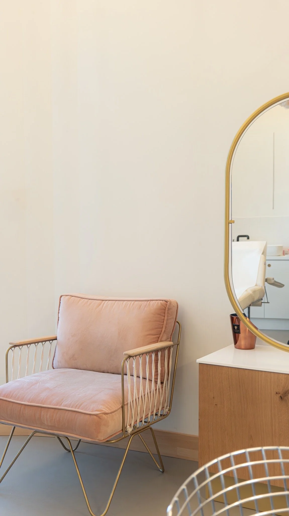 Un fauteuil en velours rose pâle avec une structure en métal doré, placé contre un mur blanc, à côté d'une petite table en bois avec un grand miroir ovale avec un cadre doré, dans une pièce moderne.