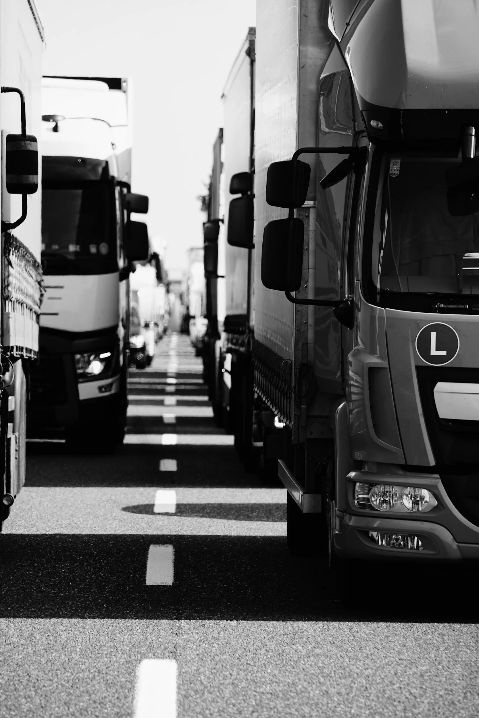 Black and white photo of trucks parked closely together on a city street, with visible side mirrors and a white dashed line dividing the lanes in the road.