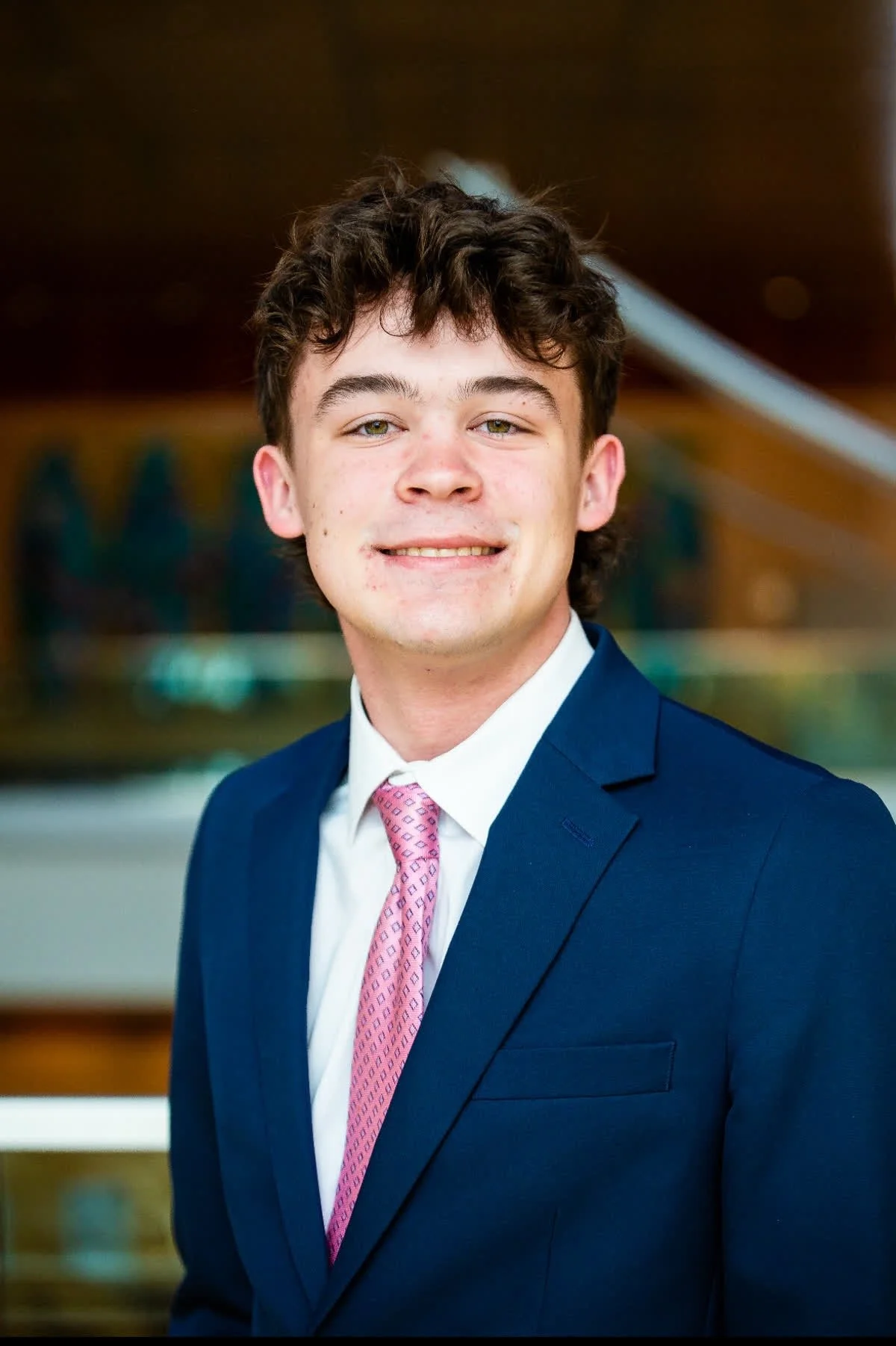 A young man with brown curly hair wearing a navy blue suit, white shirt, and pink patterned tie, smiling in front of a blurred background.