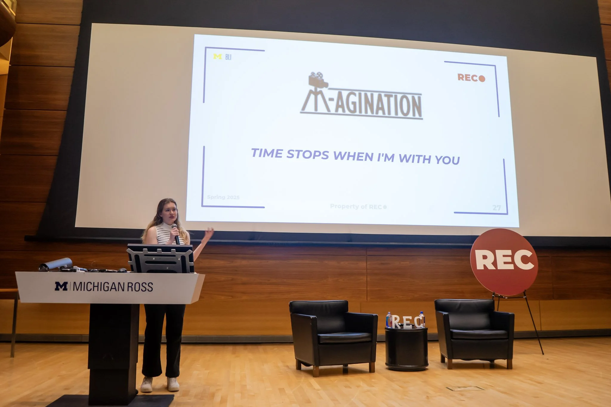 A young woman giving a presentation at Michigan Ross, standing behind a podium with a microphone and gesturing with her hand. Behind her, a large screen displays a slide titled 'Agination' with the subtitle 'Time stops when I'm with you', and logos of Michigan Ross and REC. There are two black chairs and a small table with water bottles and a red REC sign on the stage.