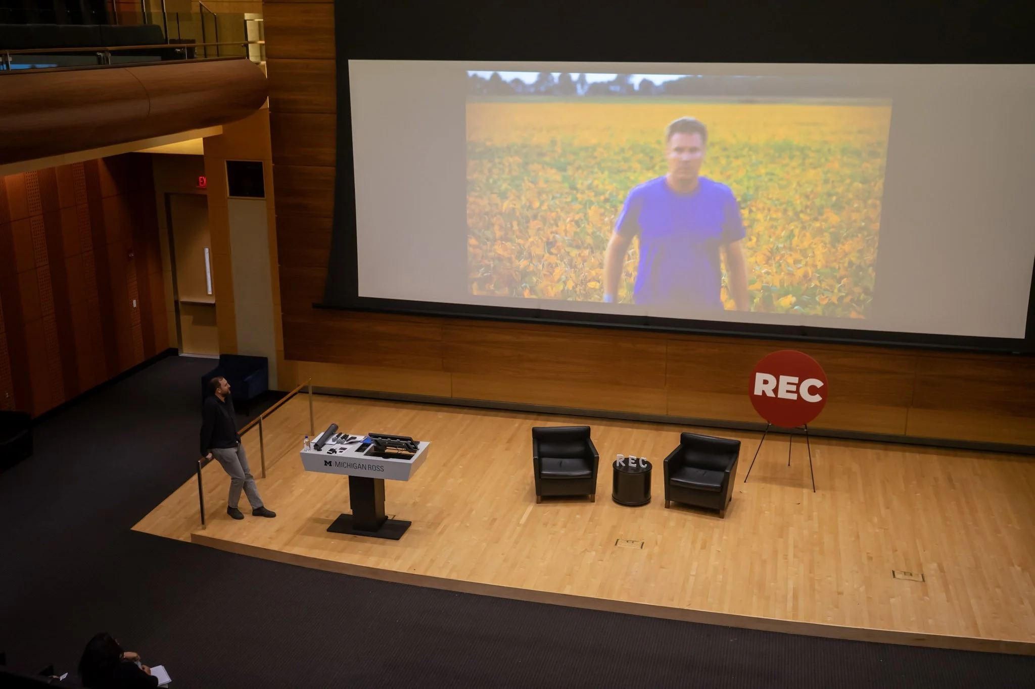 A large auditorium stage with a big screen showing a man standing in a field of yellow flowers. On the stage, there are two black chairs, a small table, and a large red sign with the word 'REC'. A woman stands on the left side near a podium labeled 'Michigan Ross' looking towards the screen.