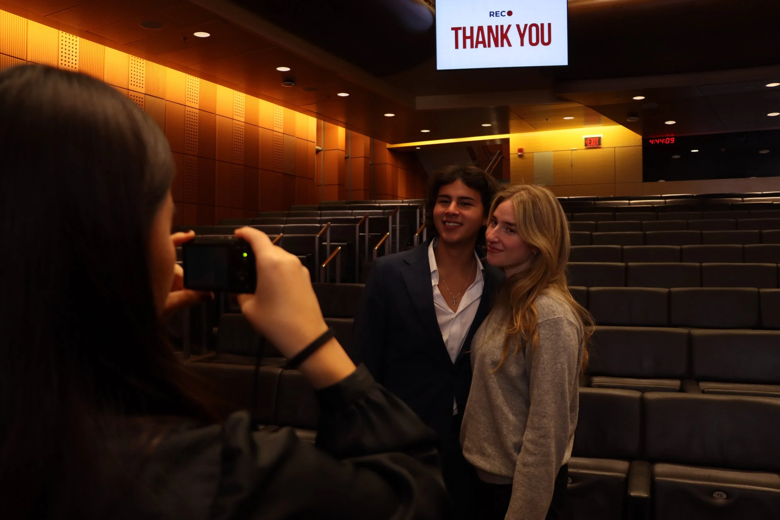 Two women posing for a photo in an auditorium with empty seats. A person in the foreground is taking their picture with a digital camera, and a screen above them displays "REC THANK YOU" in red letters.