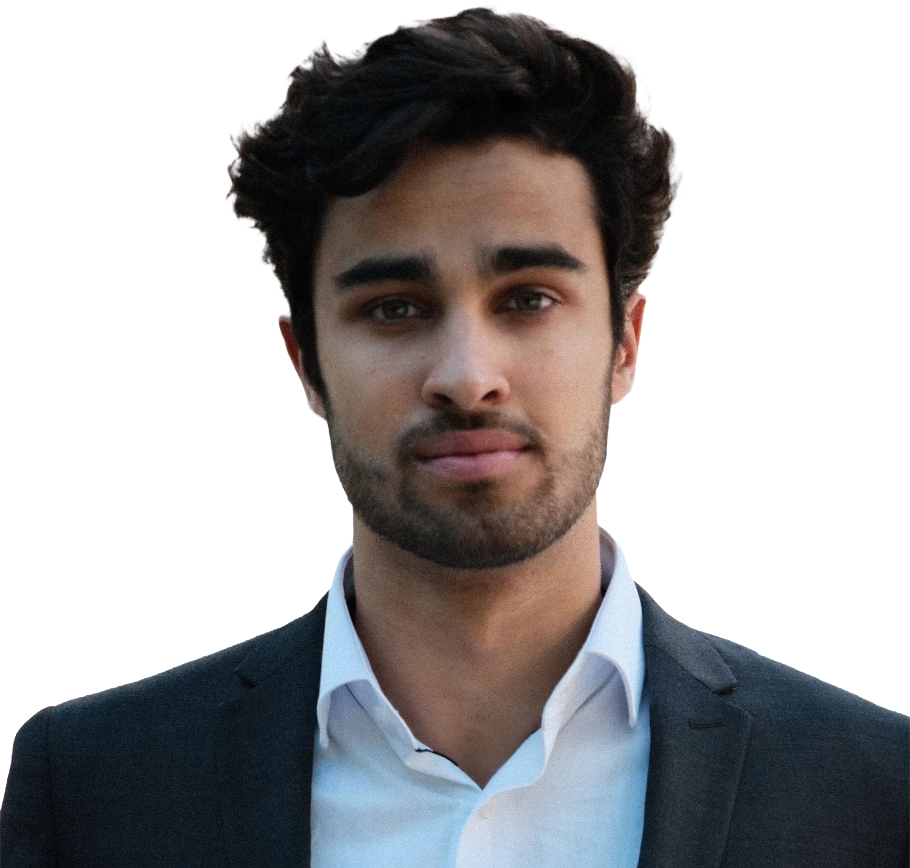 A young man with dark wavy hair, wearing a dark suit and white collared shirt, standing outdoors with a neutral expression.