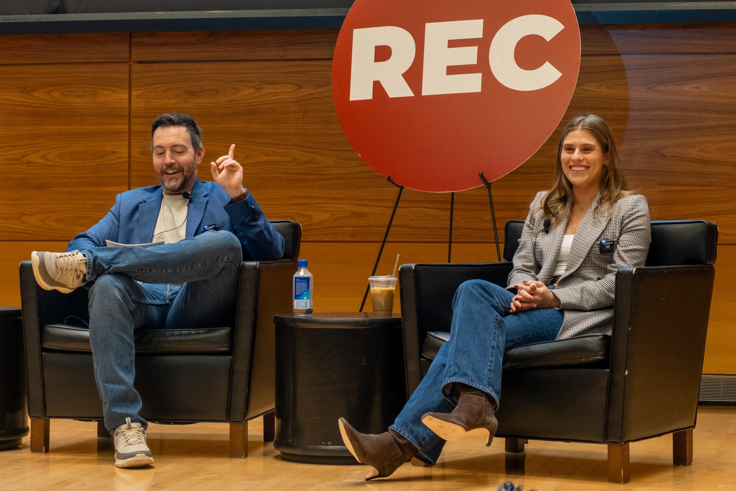 Two people seated in black armchairs on stage during a discussion or interview. The man on the left, with short dark hair and a beard, is wearing a blue blazer, white shirt, jeans, and sneakers. He appears to be speaking or laughing, with one hand raised. The woman on the right, with long light brown hair, is wearing a checkered blazer, white top, jeans, and brown heeled boots. She is smiling. Between them is a small table with a bottle of water, a coffee cup, and a straw. In the background, there is a large red circle with the word "REC" on it, indicating recording.