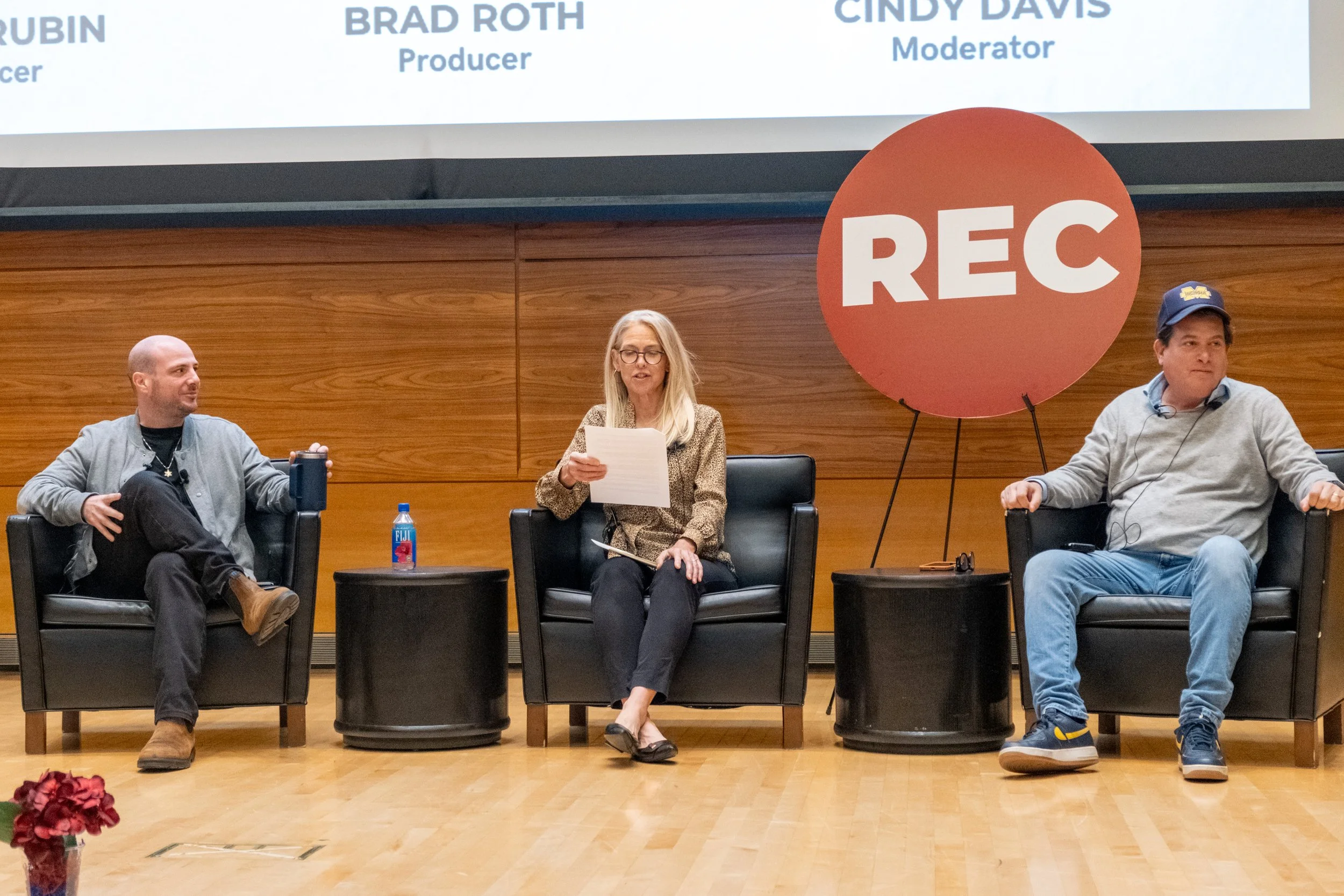 Three people seated on a panel at a recorded event, with two men on either side and a woman in the middle. The woman is reading from a sheet of paper. There is a large recording sign ('REC') on a stand next to the woman. A large screen behind them displays names and titles, including Brad Roth as producer and Cindy Davis as moderator.