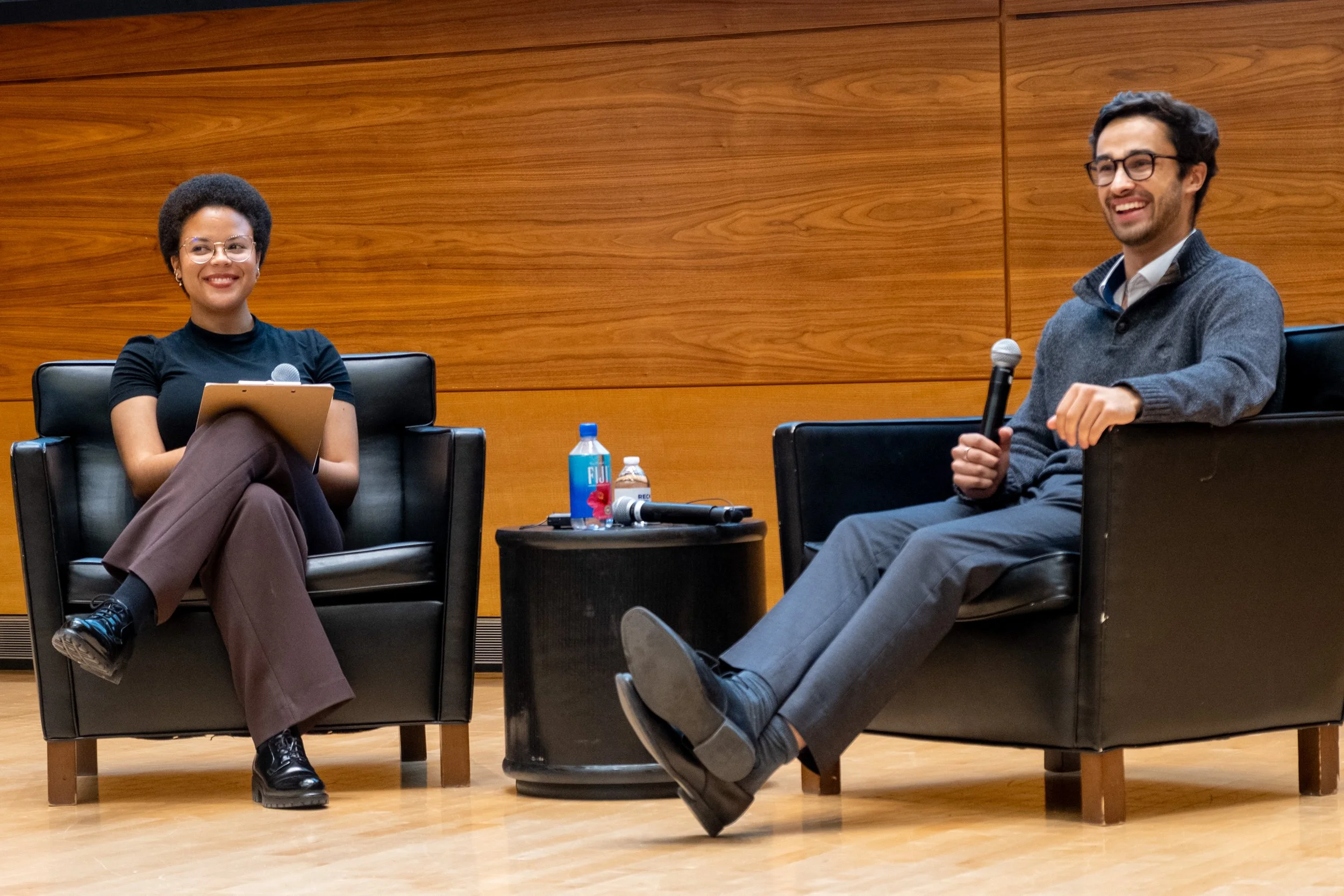 Two people seated in black armchairs having a discussion. The woman on the left has short curly hair, glasses, and is holding a notepad and pen. The man on the right has dark hair, glasses, and is holding a microphone. There is a small table between them with water bottles, a soda can, a microphone, and a coffee mug. The background is a wooden-paneled wall.