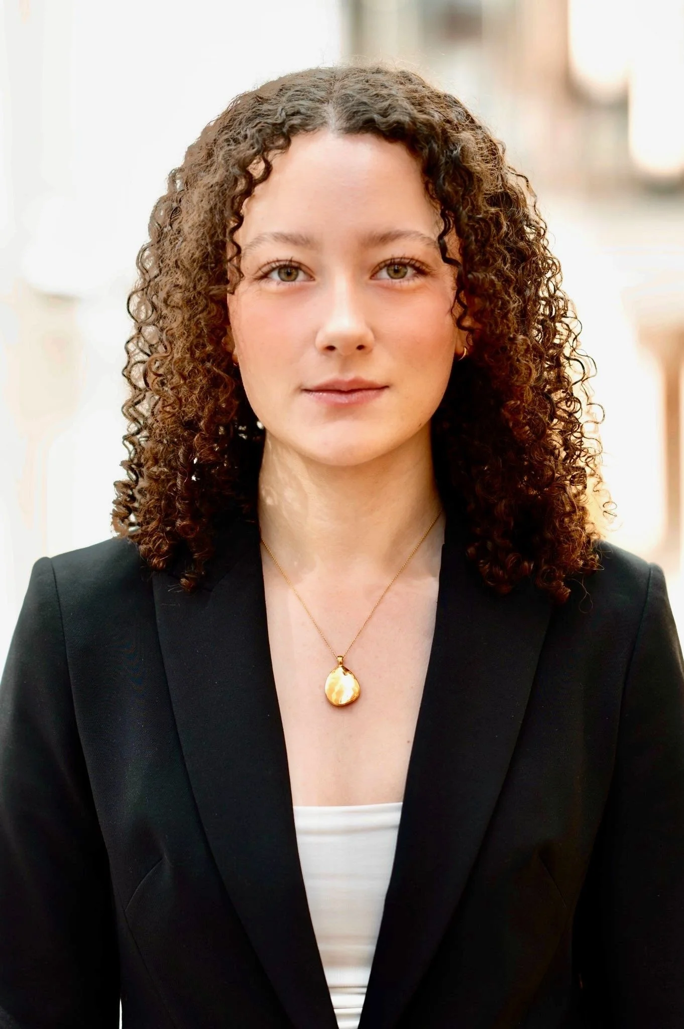 A woman with curly brown hair and light skin wearing a black blazer, a white top, and a gold pendant necklace.