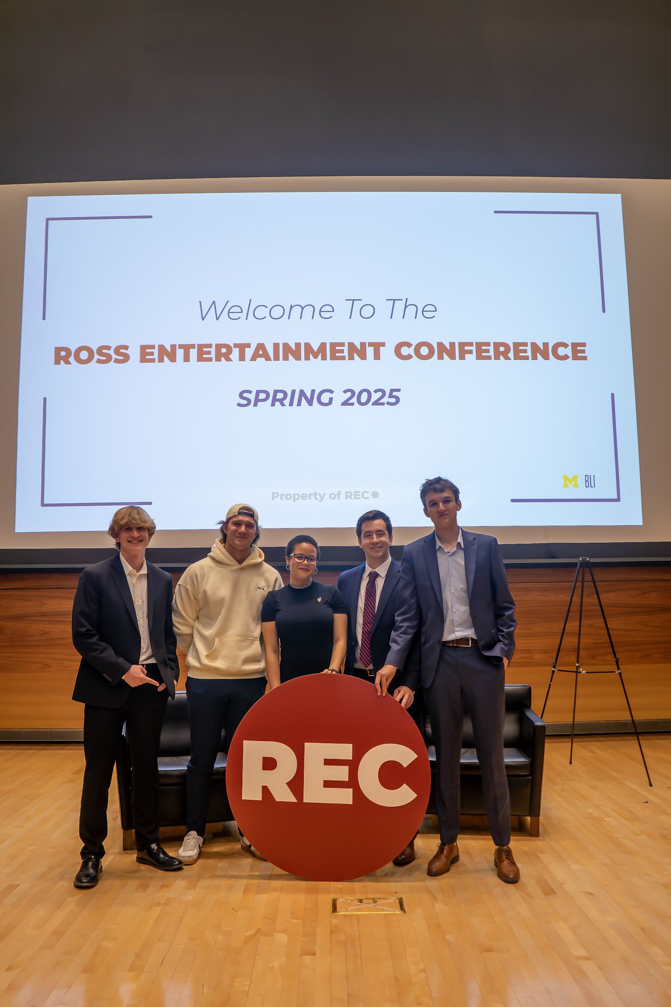 Group of five young people standing in front of a large screen that reads "Welcome to the ROSS ENTERTAINMENT CONFERENCE SPRING 2025," with a red circular sign with white letters "REC" in front of them.