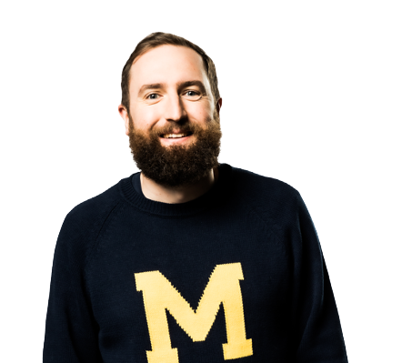 A man with a beard wearing a black sweater with a yellow 'M' on it, smiling, against a black background.