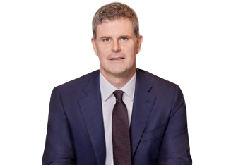 A man wearing a dark blue suit, white shirt, and dark tie, sitting against a plain background.