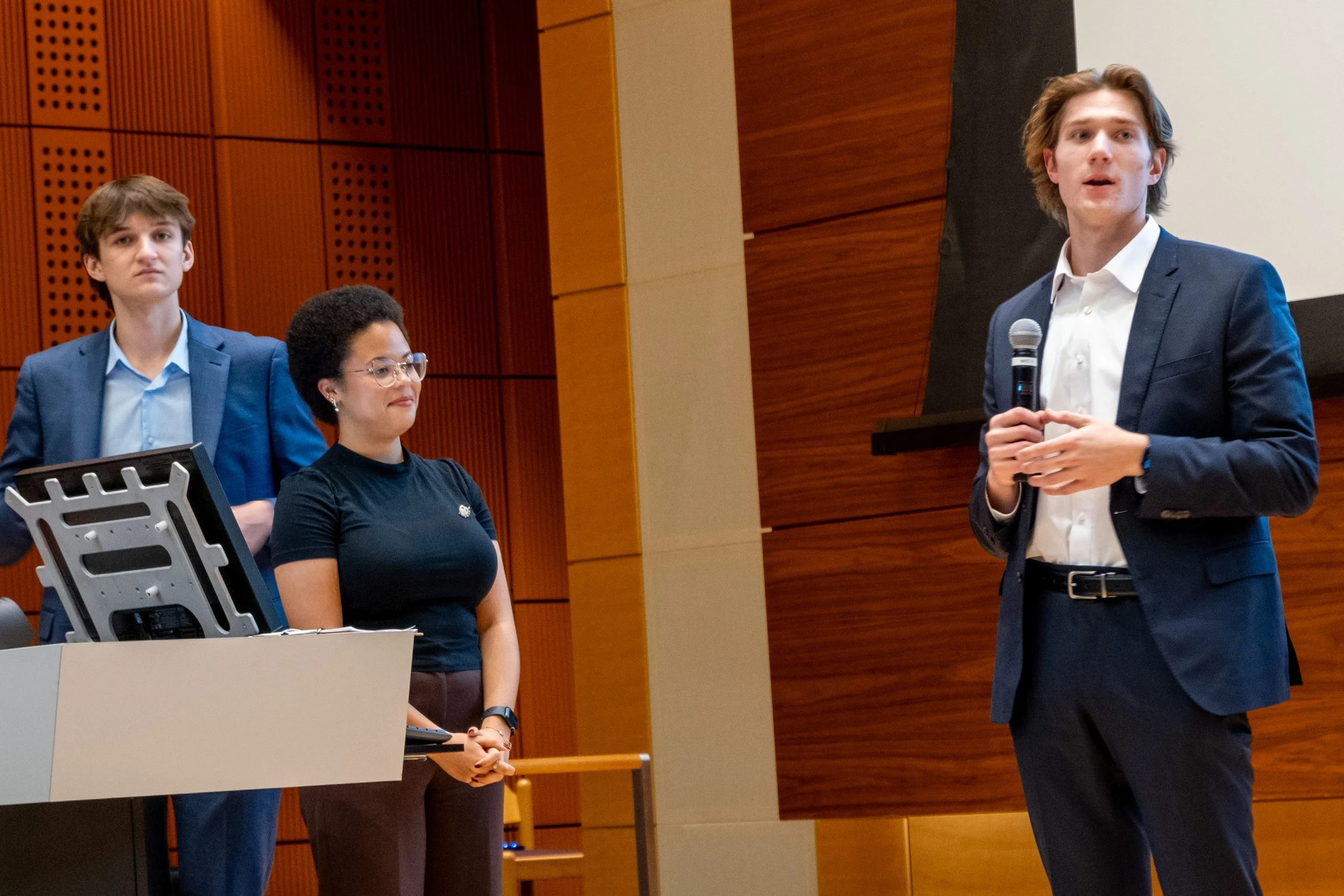 A man in a dark blue suit is speaking into a microphone in front of an audience during a presentation. There are three other people standing behind him, two men and one woman, all dressed professionally. The setting is an auditorium with wooden walls.