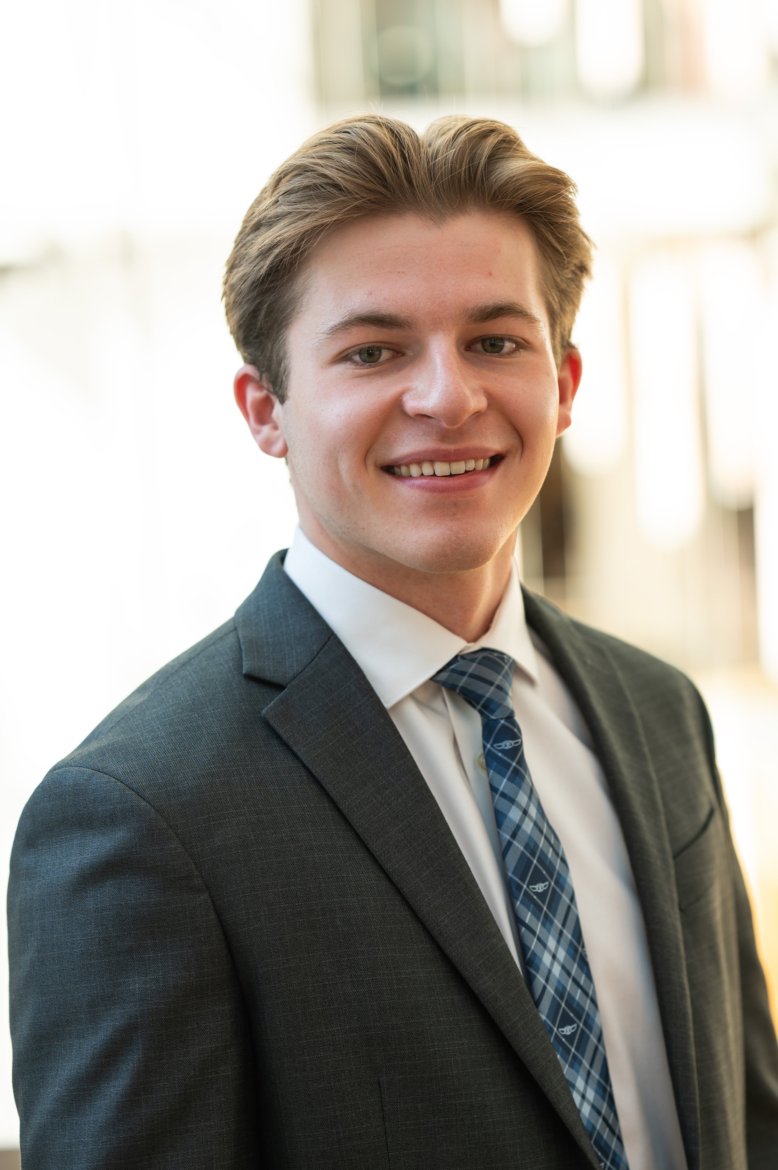Young man in a dark suit, white shirt, and plaid tie, smiling in a professional setting with sunlight in the background.