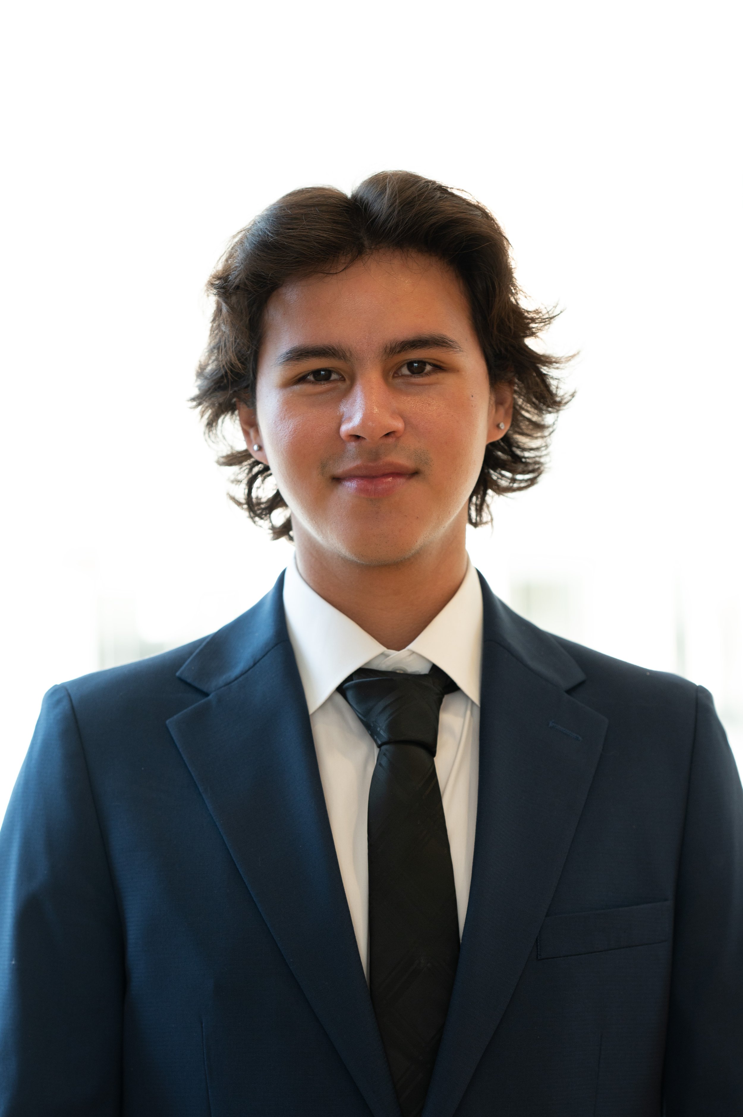 Young man in a navy suit and tie, smiling softly, with a blurred bright background.