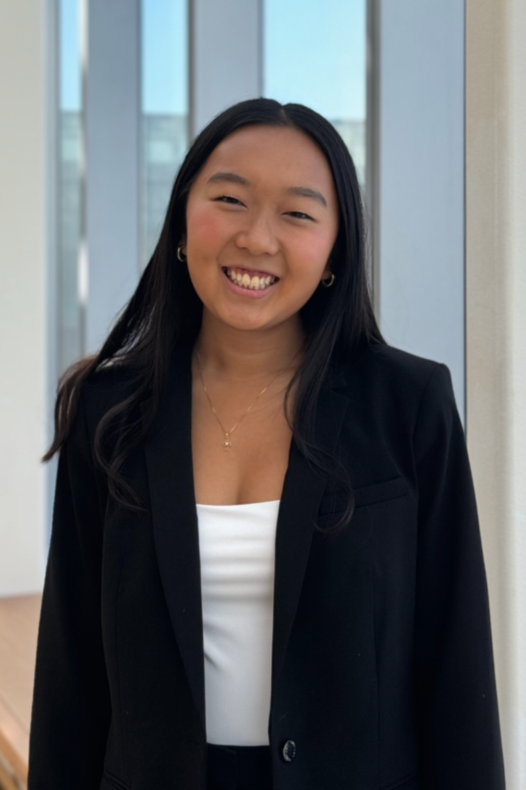 A young woman with long black hair smiling, wearing a black blazer and white top, standing indoors near a window.