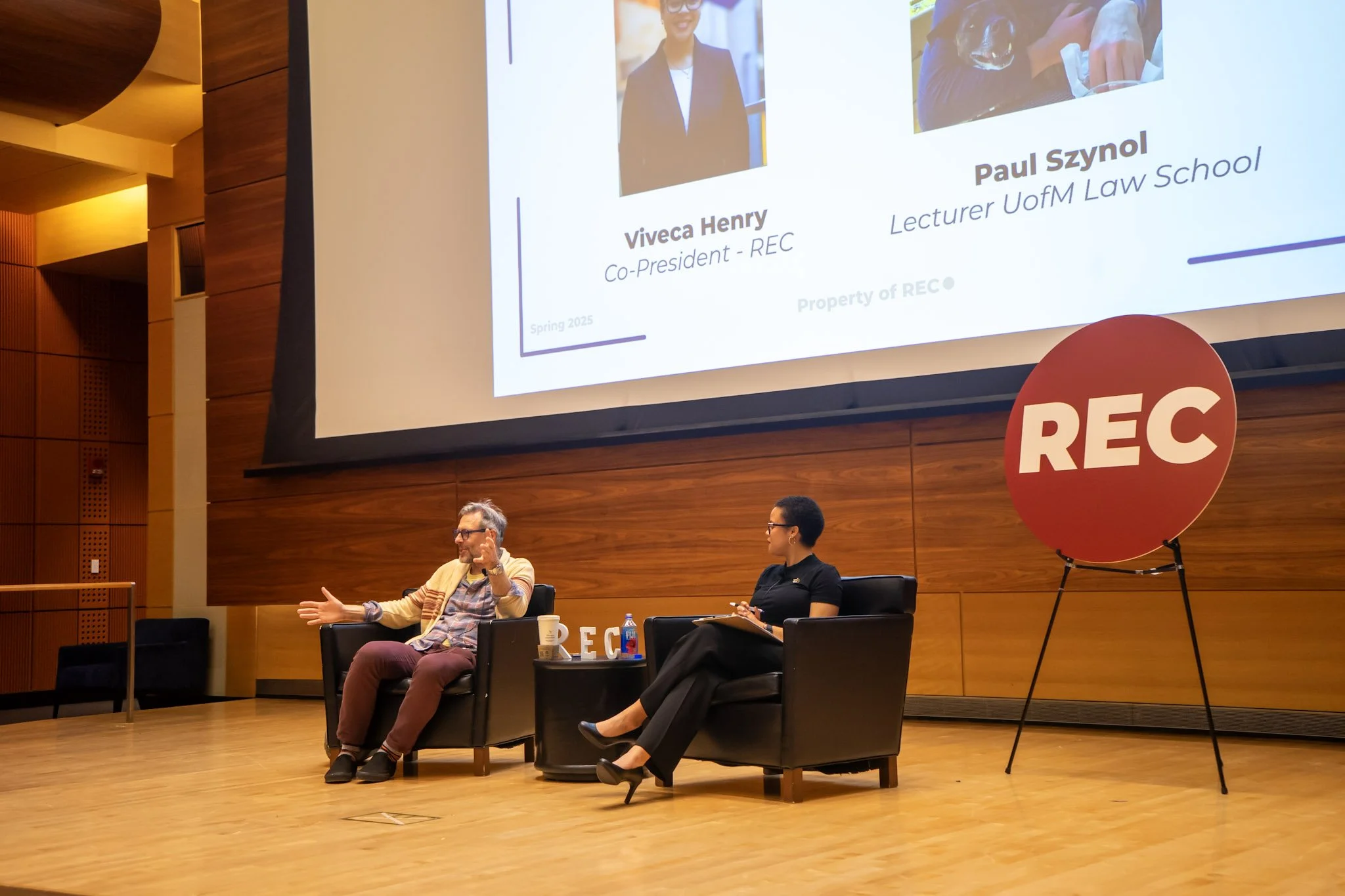 Two people seated on a stage engaged in a discussion, with a large screen behind them displaying information about Viveca Henry and Paul Szyzol. The stage has a wooden backdrop and a large red circle with the word "REC" on it, indicating recording of the event.