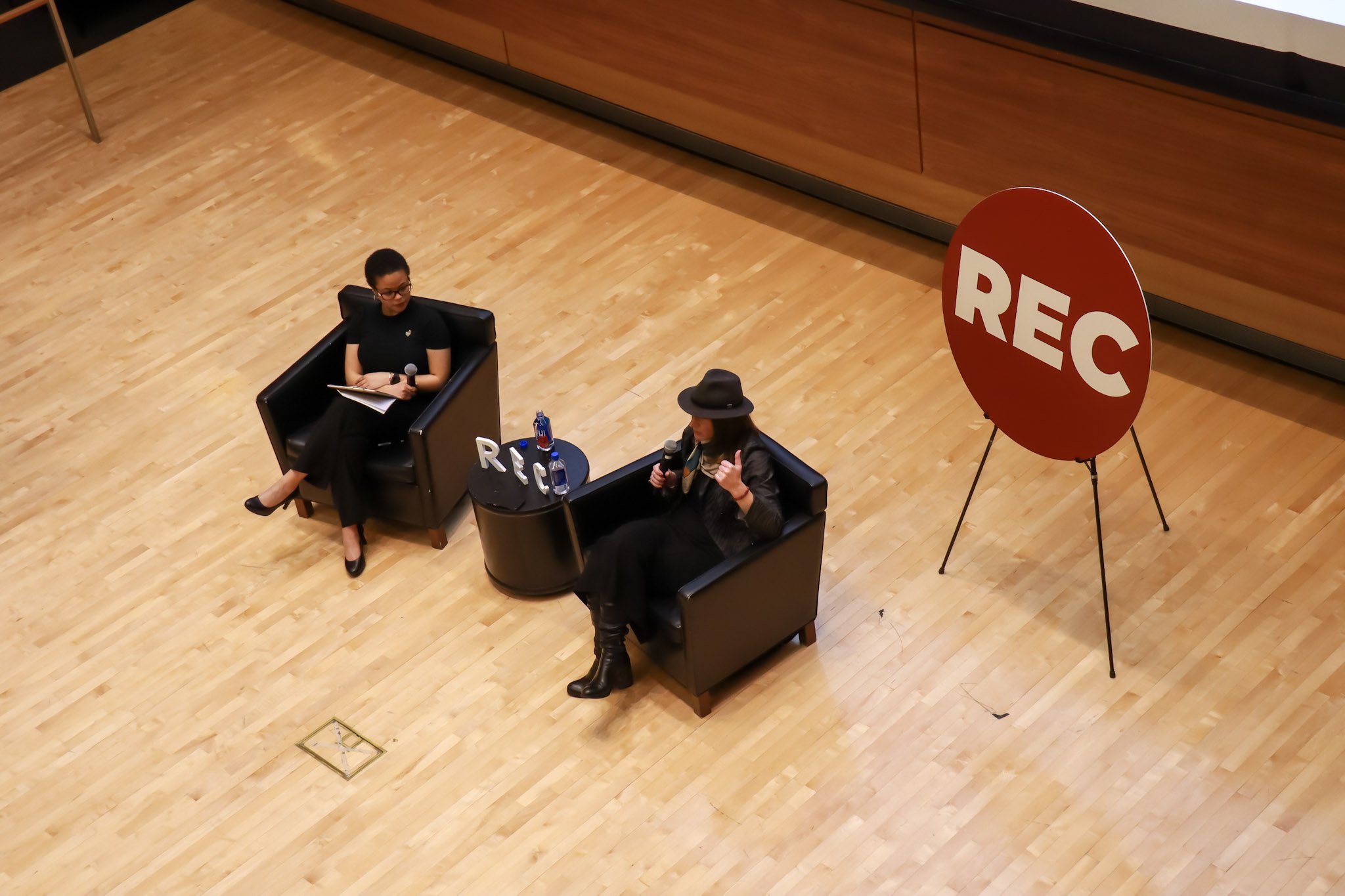 Two women sit in black armchairs on a wooden floor, engaged in a discussion with microphones. One woman wears a black hat and boots, and the other woman wears glasses and black shoes. A sign with the letters 'REC' is on a table between them, along with two water bottles. A large red circular sign with 'REC' is nearby.