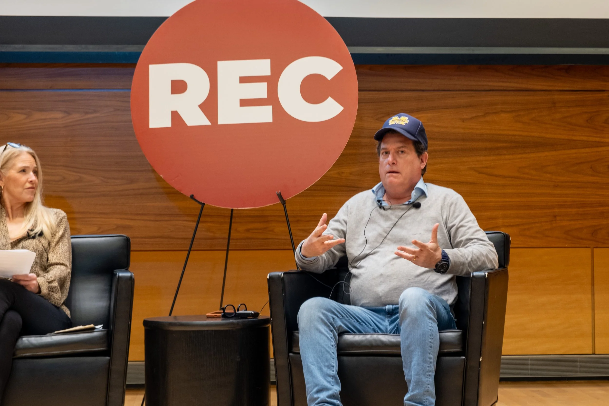 A man in a blue cap and casual clothing speaking during a panel discussion, seated in front of a large red sign with white letters that says 'REC'; a woman with long blonde hair and glasses sits to his left, holding a notebook.