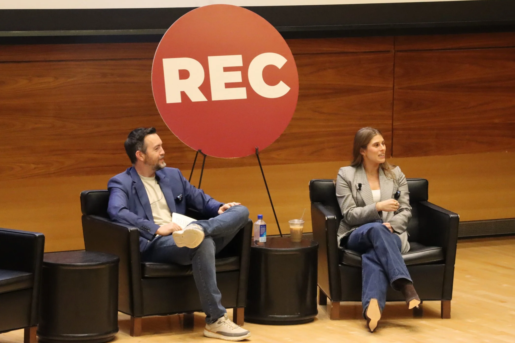 Two people sitting in black armchairs on a stage, one man and one woman, with a large red circle sign with 'REC' behind them, microphones in hand, and a table with a drink and water bottle between them.