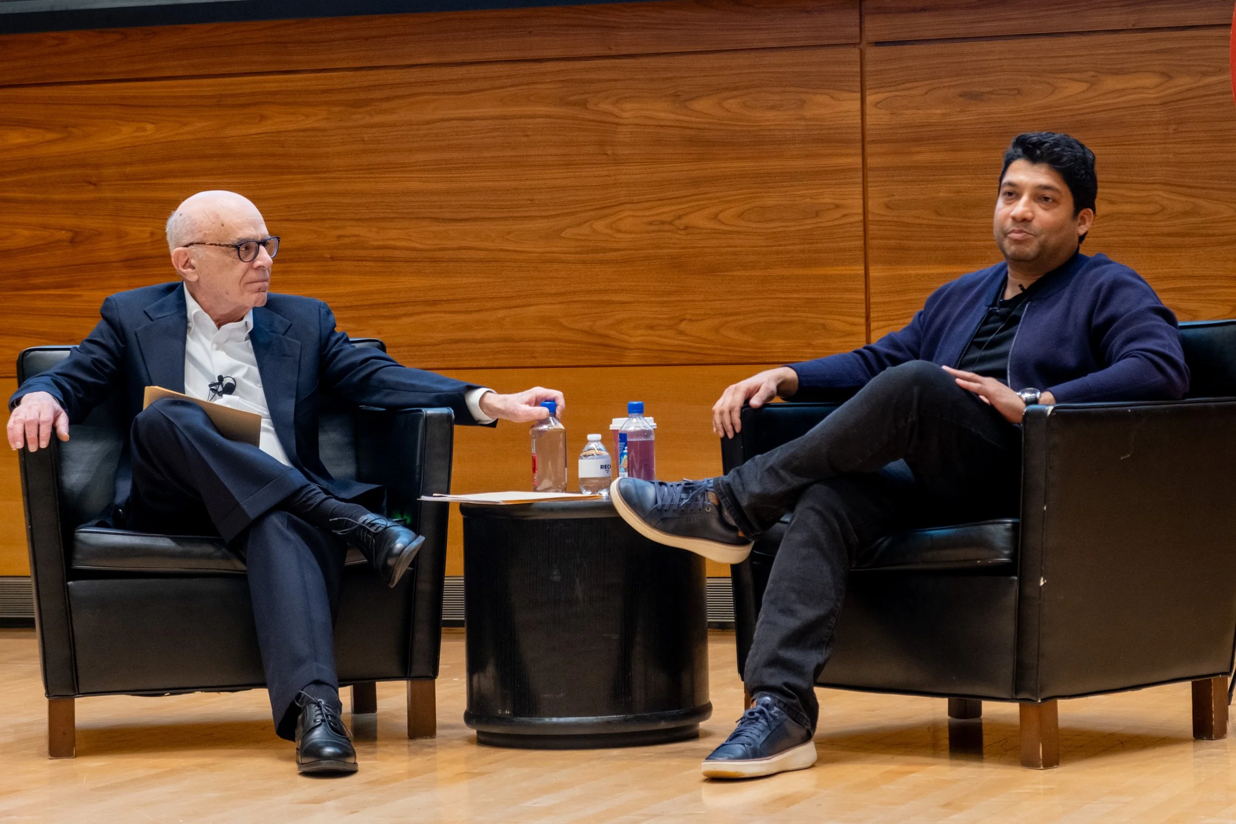 Two men are sitting in armchairs on a stage, engaged in a conversation. The man on the left is elderly, bald, wearing glasses, a navy suit, white shirt, and black shoes. The man on the right is younger, with dark hair, wearing a navy jacket, black t-shirt, and black pants. There is a small black table between them with water bottles and hand sanitizer. The background features a wooden panel wall.