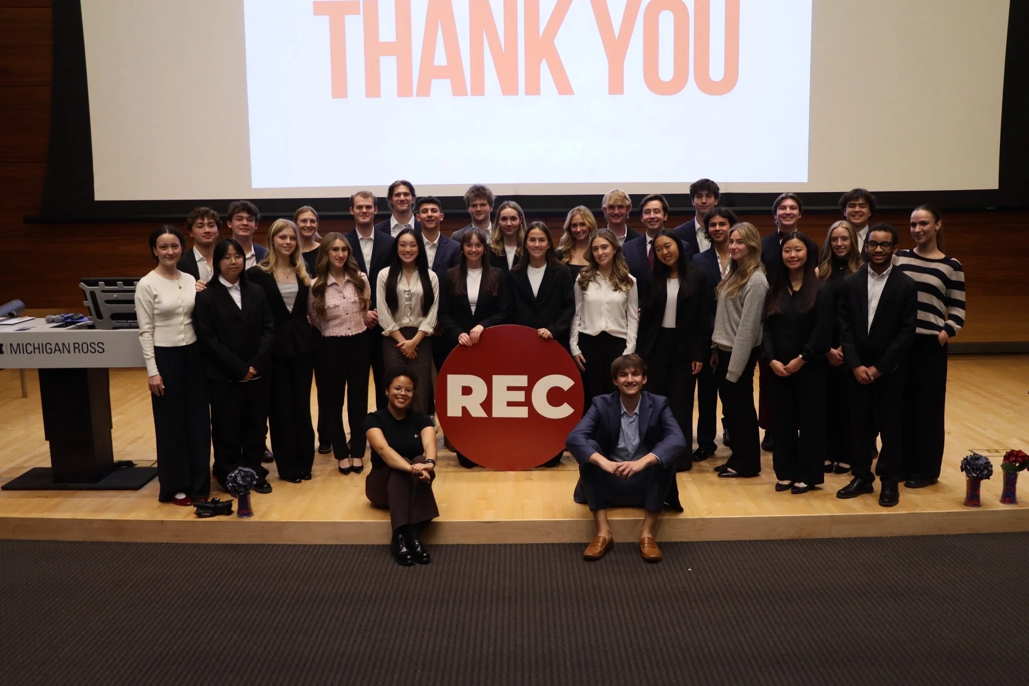 Group of people on stage celebrating with a large 'REC' sign, in front of a screen displaying 'THANK YOU', with bouquet flowers on the floor and a keyboard on the podium to the side.