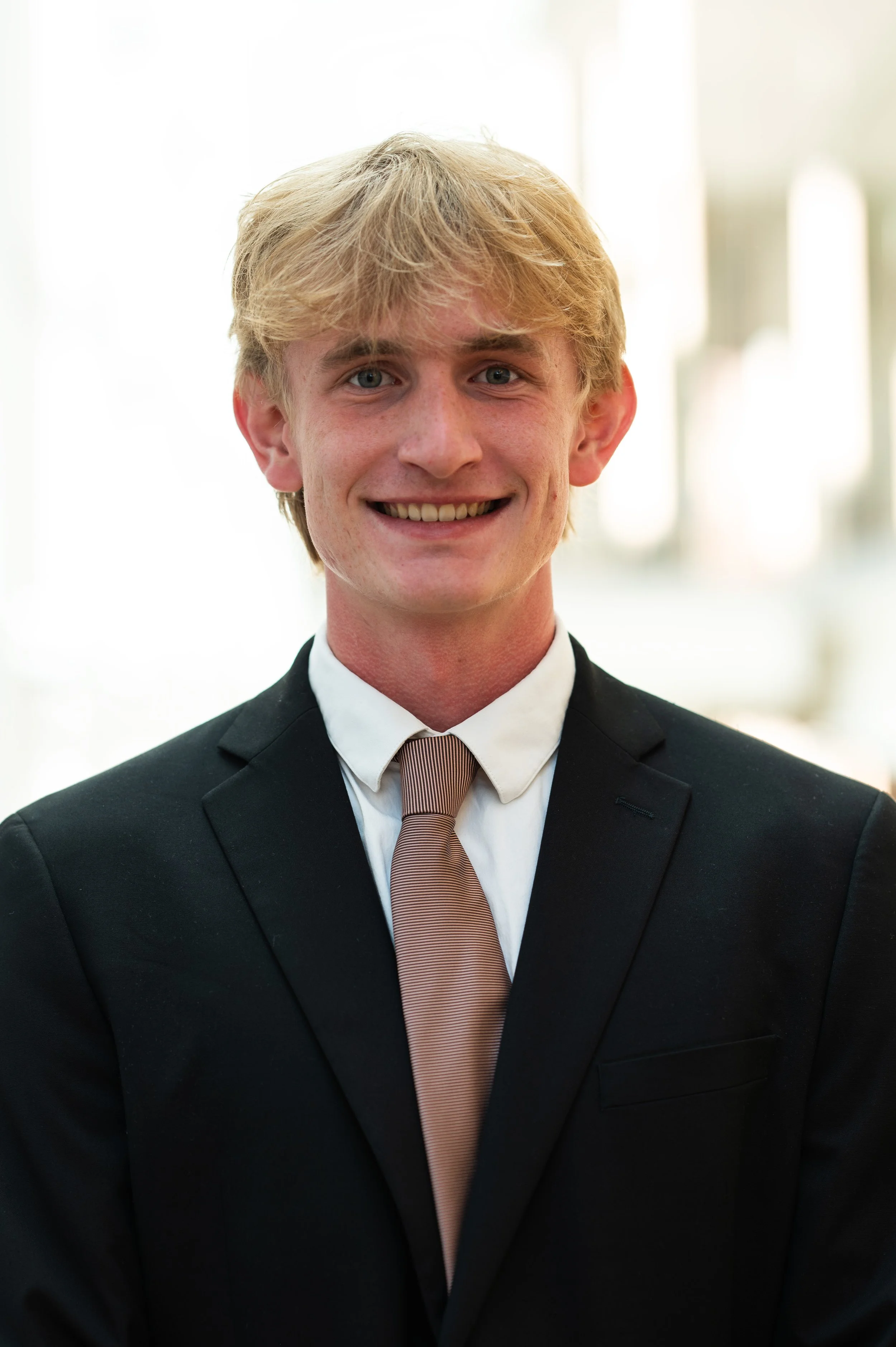 Young man with blond hair and blue eyes wearing a black suit, white shirt, and a light brown tie smiling at the camera, standing indoors with blurred windows in the background.