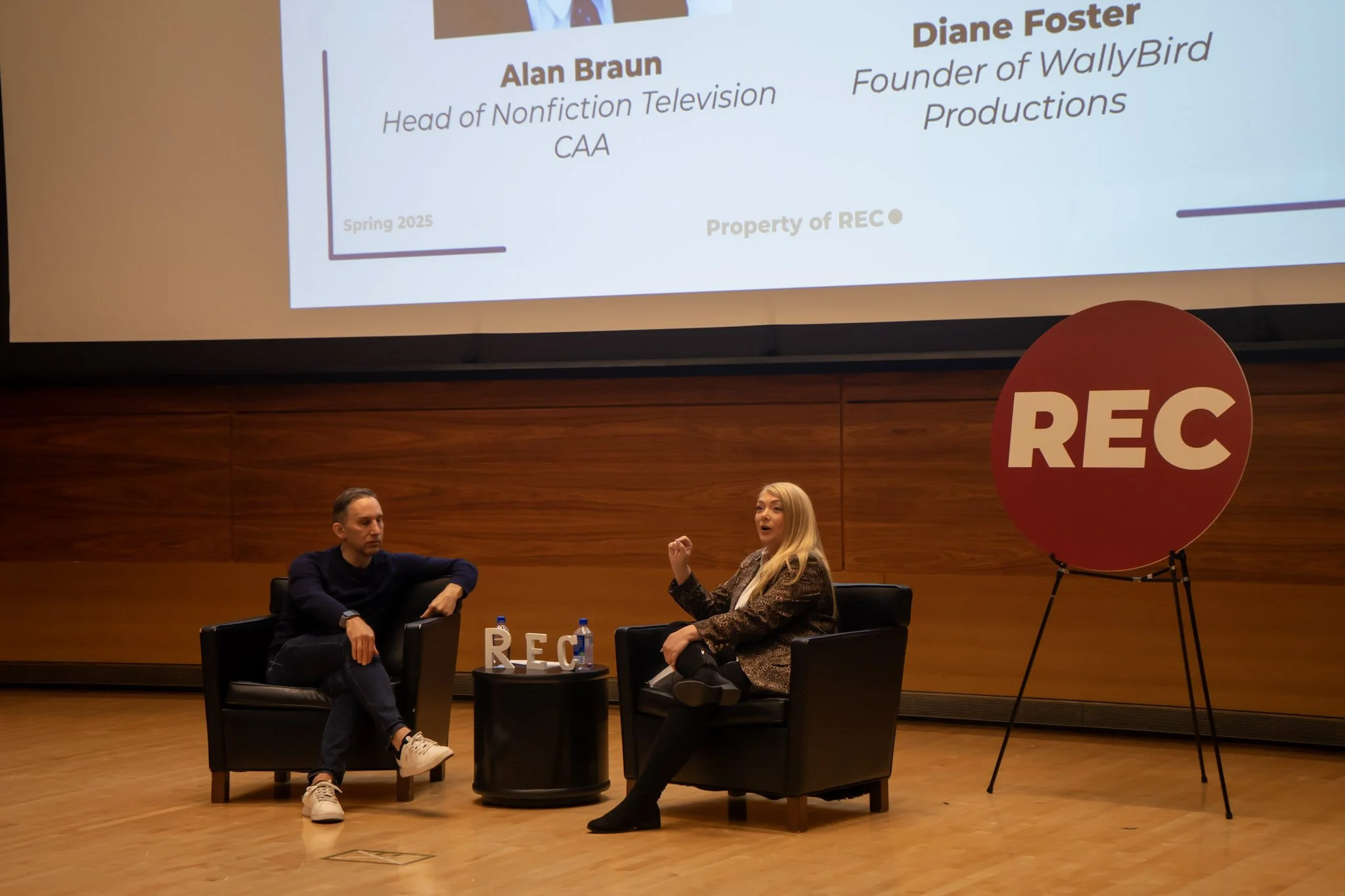 Two people sitting in armchairs on a stage with a large screen behind them displaying names and titles; a woman speaking and gesturing, a large red REC sign on stand nearby, and small round table with two water bottles and decorative letters 'REC'.