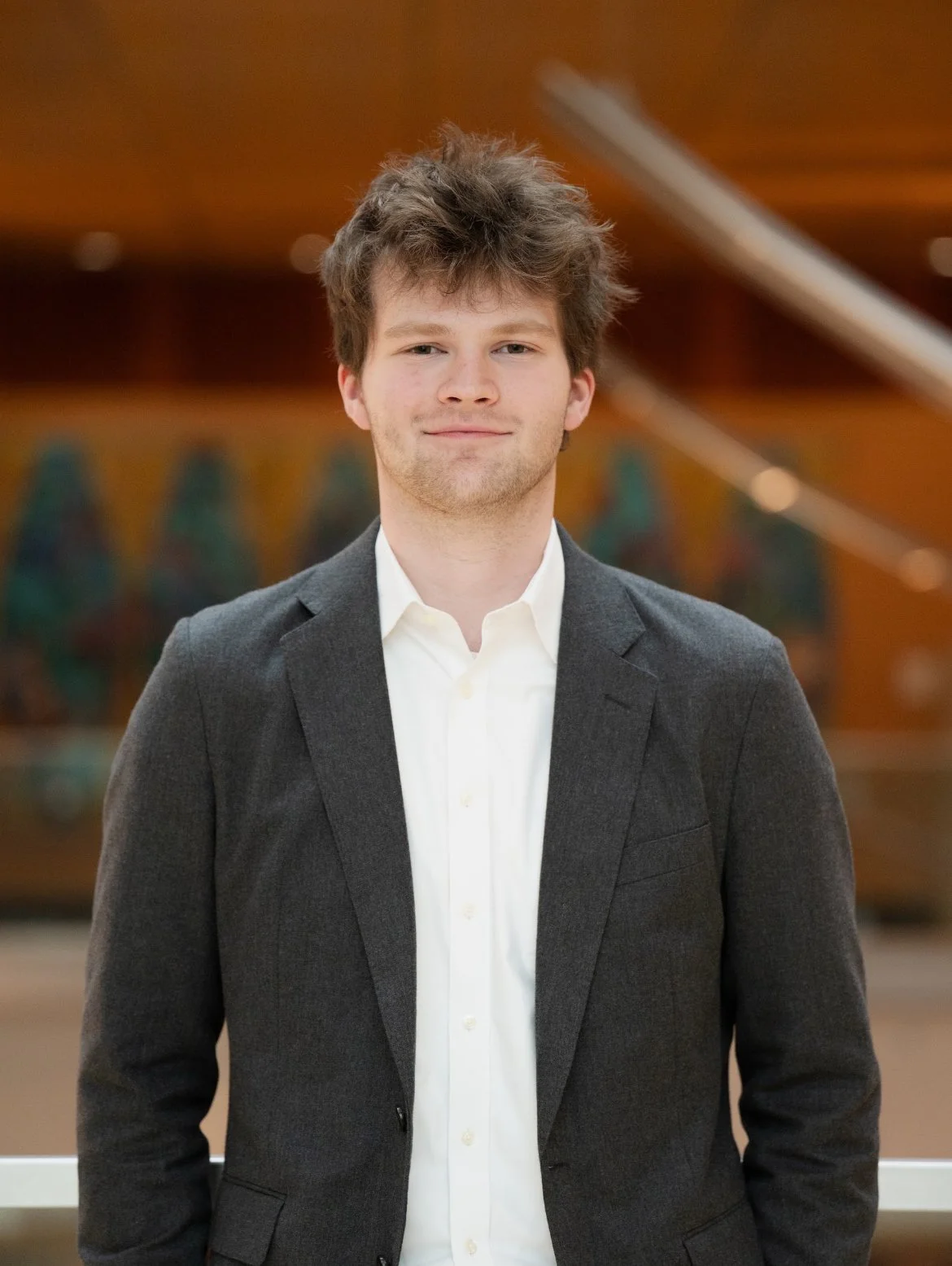 A young man with curly brown hair, wearing a dark gray blazer and white shirt, standing indoors with a blurred background of a wooden structure and artwork.