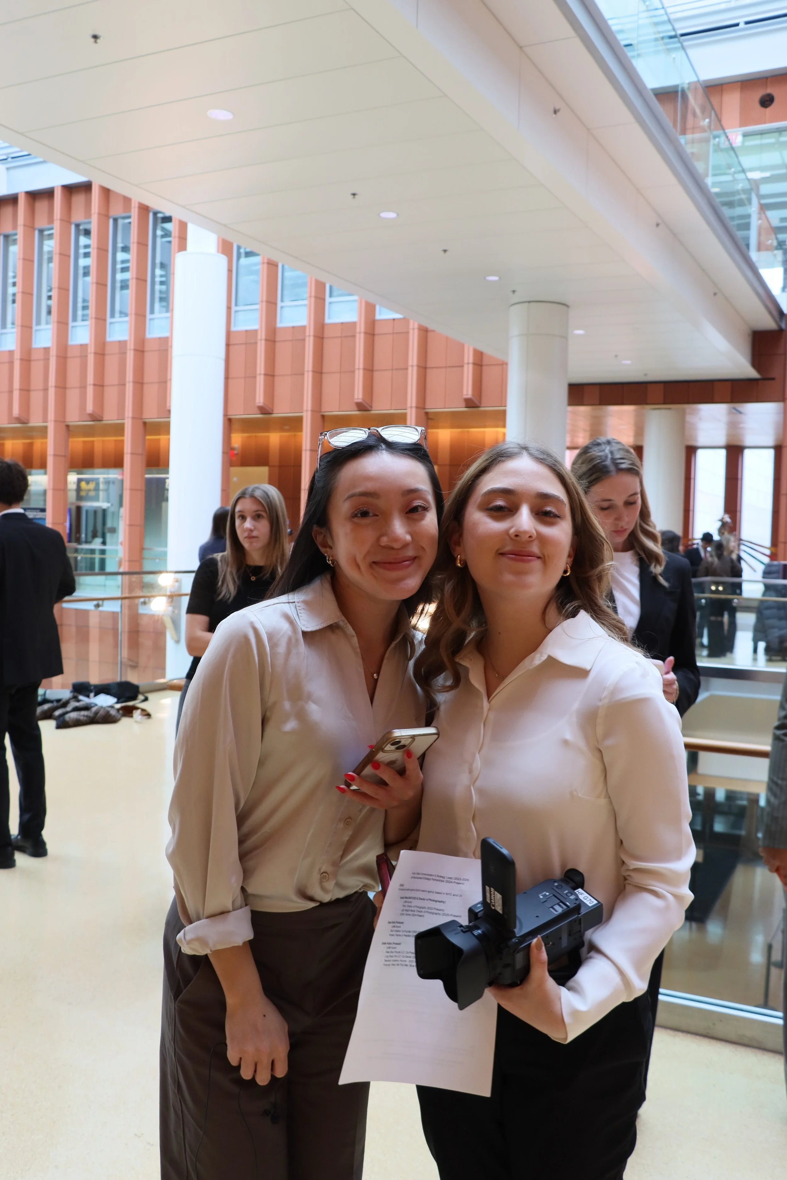 Two women smiling at the camera, holding a document and a camera, in a modern indoor public space with large windows and other people in the background.