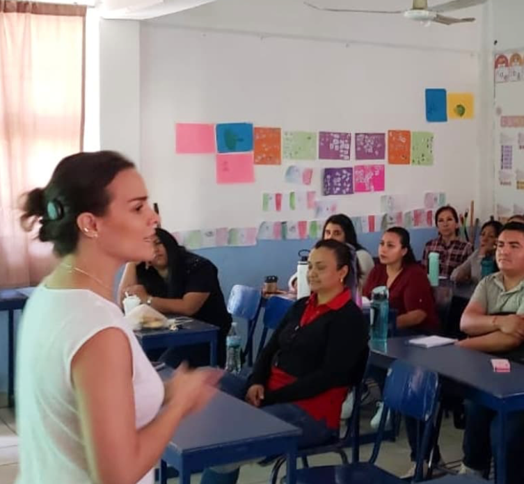 Montse, una mujer que se encuentra frente a un grupo de docentes de un Colegio dentro de un aula, donde imparte una charla sobre cómo potenciar y mejorar apoyos para la comunicación de los alumnos dentro del aula de clases.